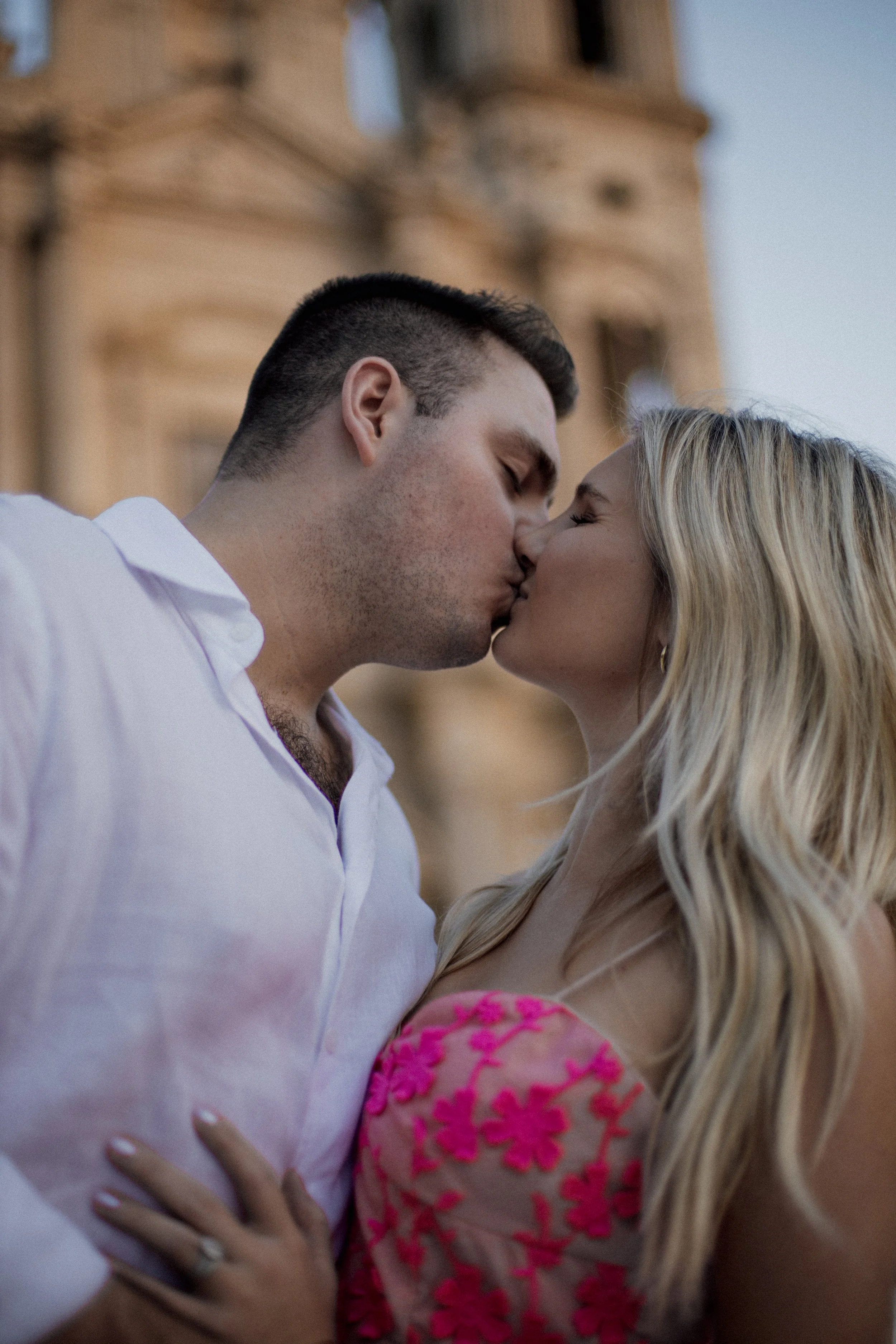 A couple sharing a kiss outdoors with an ornate building in the background.