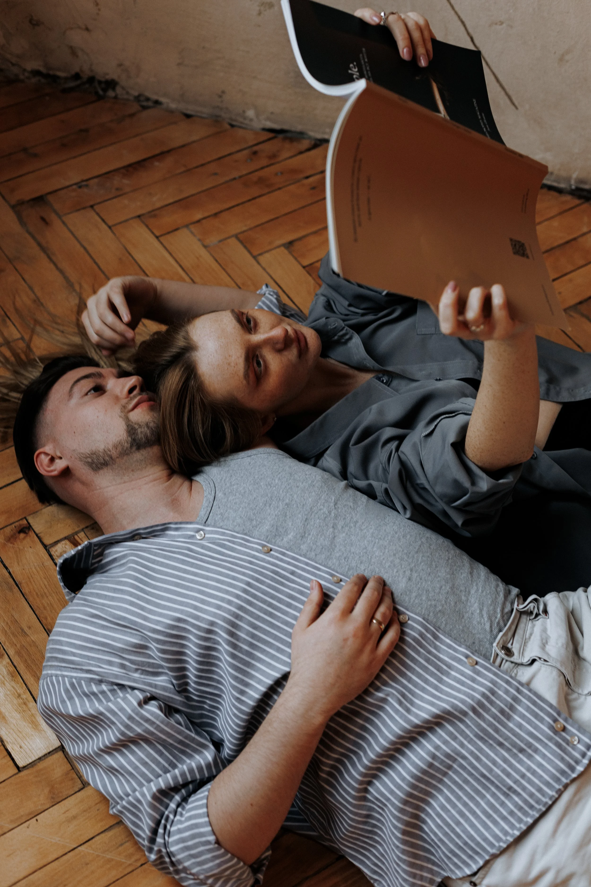 A man and woman lying on a hardwood floor, sharing a relaxing moment while reading a magazine.