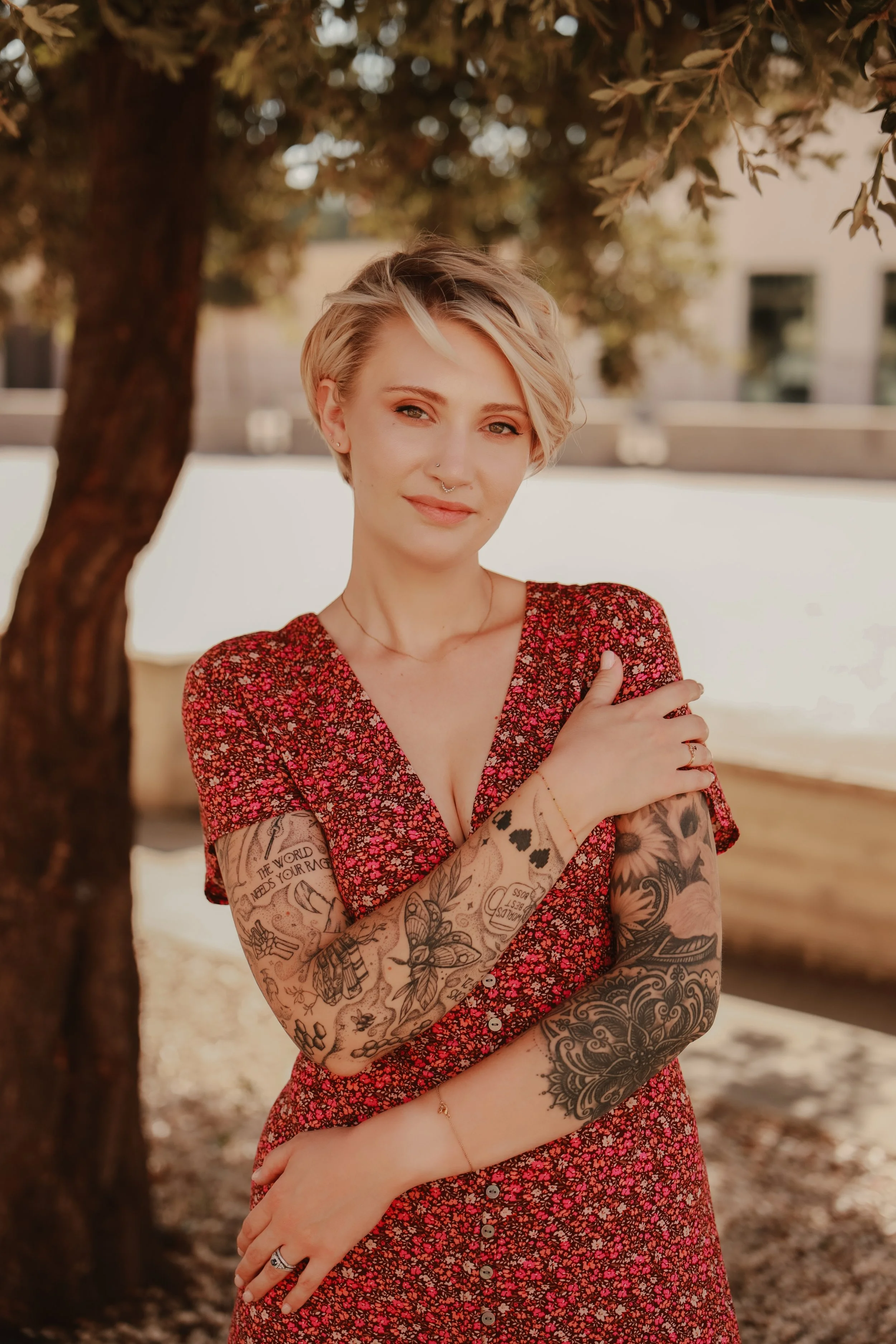 A woman with short blonde hair and tattoos on her arms stands outdoors under a tree. She wears a red floral dress and smiles gently.