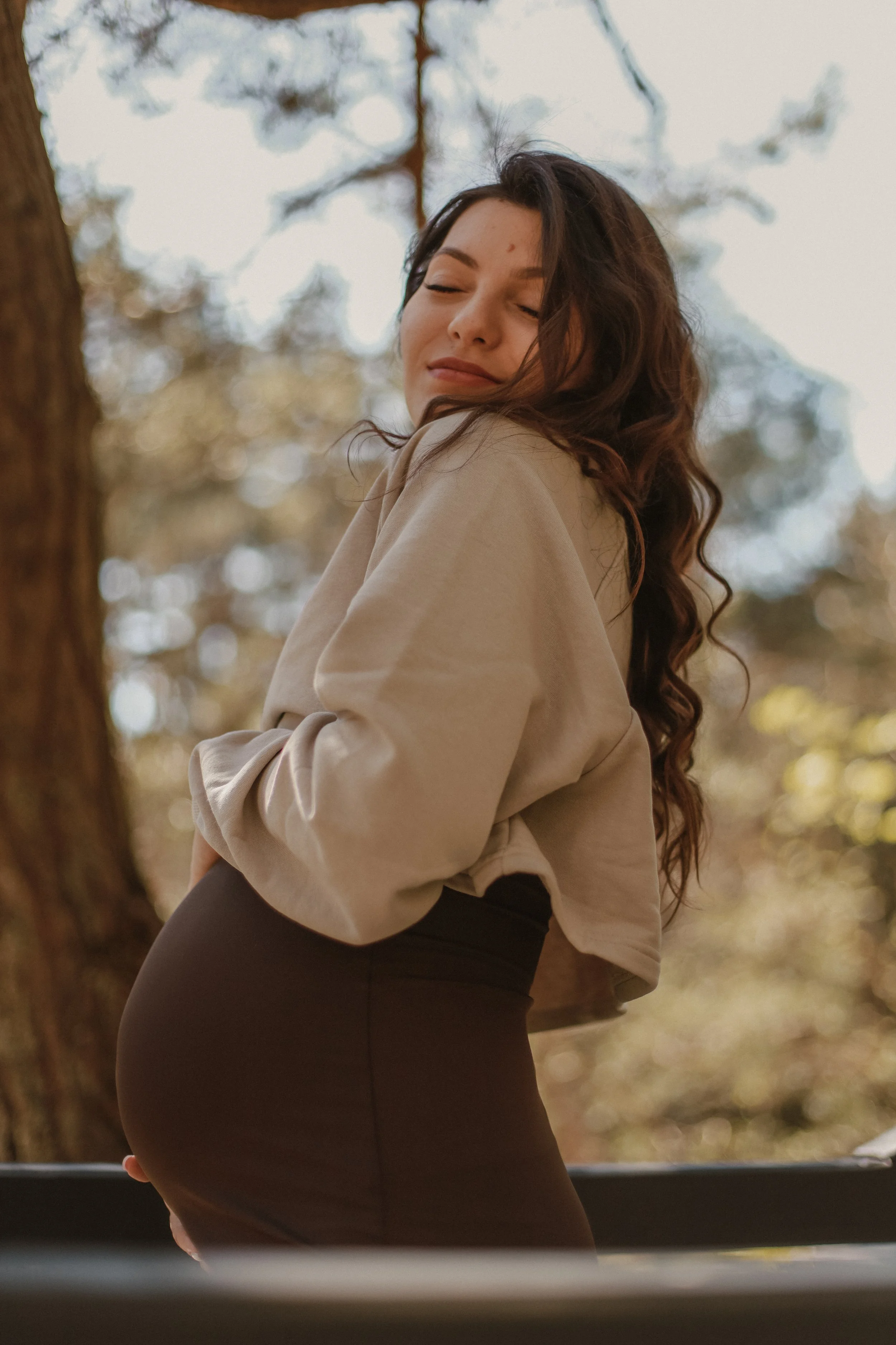 A woman with long, wavy brown hair standing outdoors in a forest, smiling with eyes closed, wearing a beige jacket and dark pants, with a background of trees and sunlight filtering through