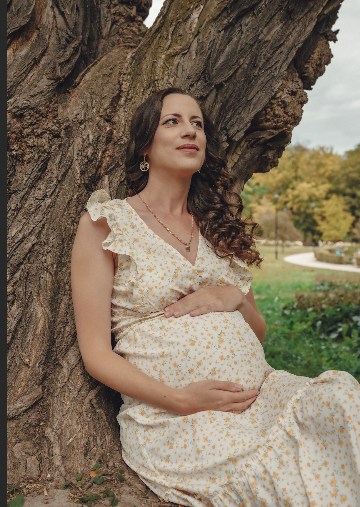 A pregnant woman in a floral dress sitting against a large tree outdoors during daytime, looking thoughtfully into the distance.