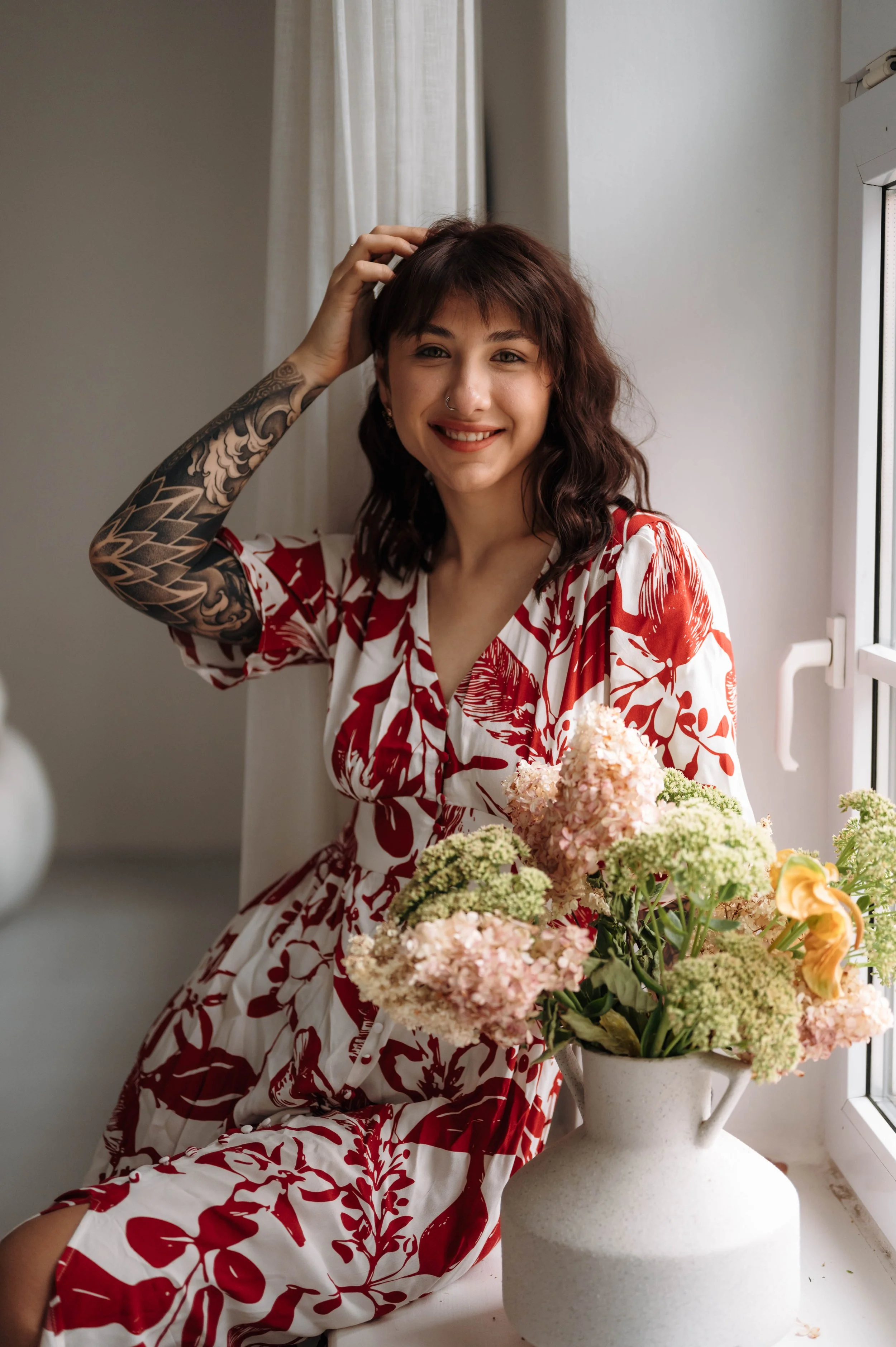 Woman with tattoos on her arm, smiling, sitting near a window with a large bouquet of pink and green flowers in a white vase.