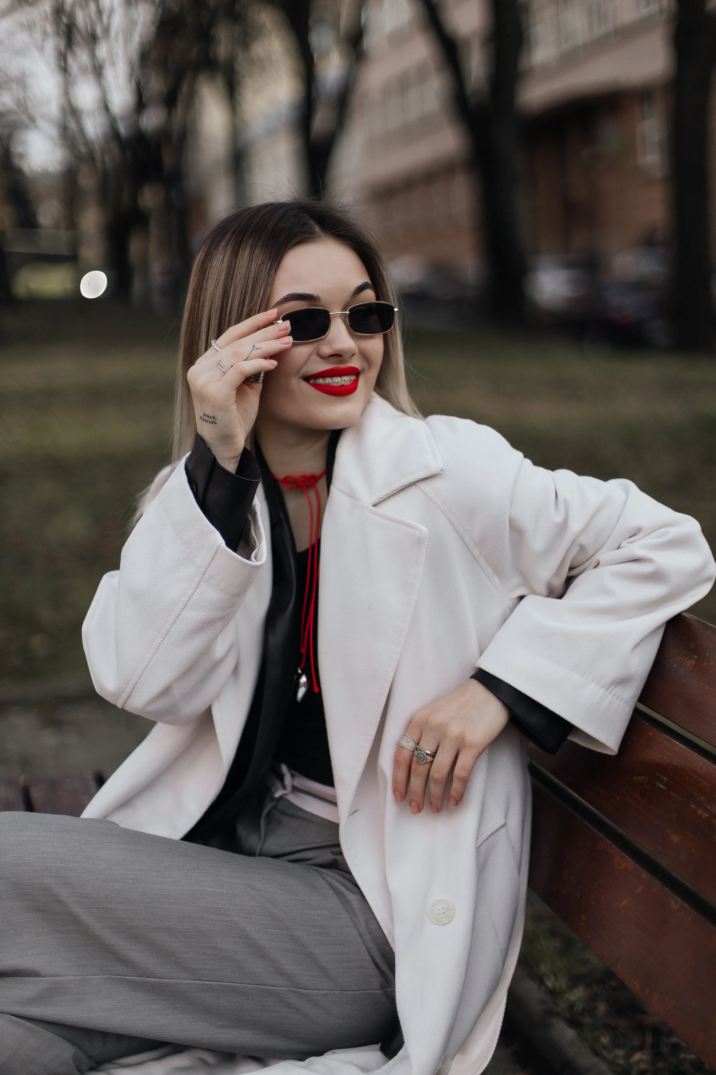 A woman sitting on a park bench, wearing sunglasses, a white blazer, gray pants, red lipstick, and smiling.