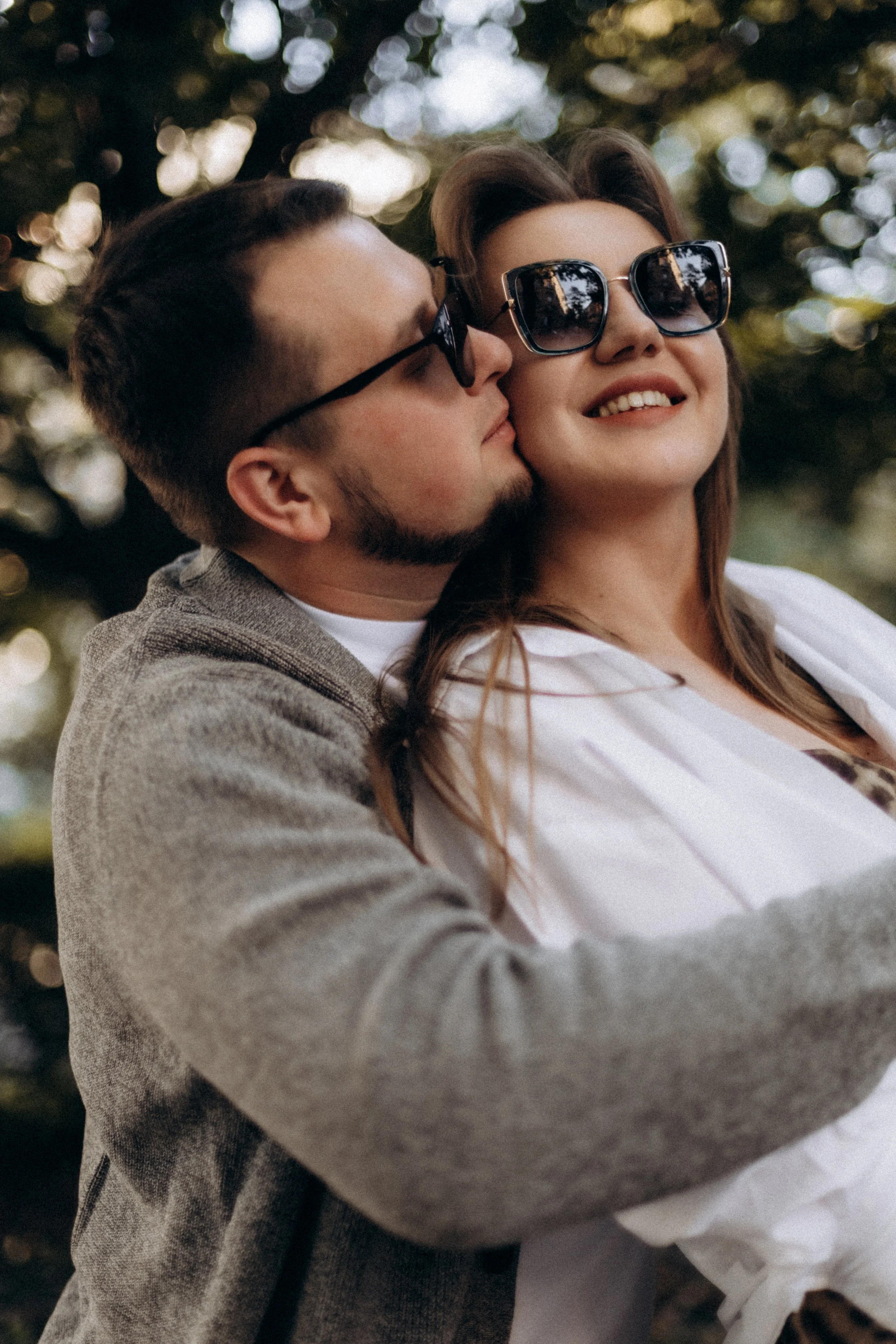A man and woman wearing sunglasses, embracing outdoors with trees and sunlight in the background.