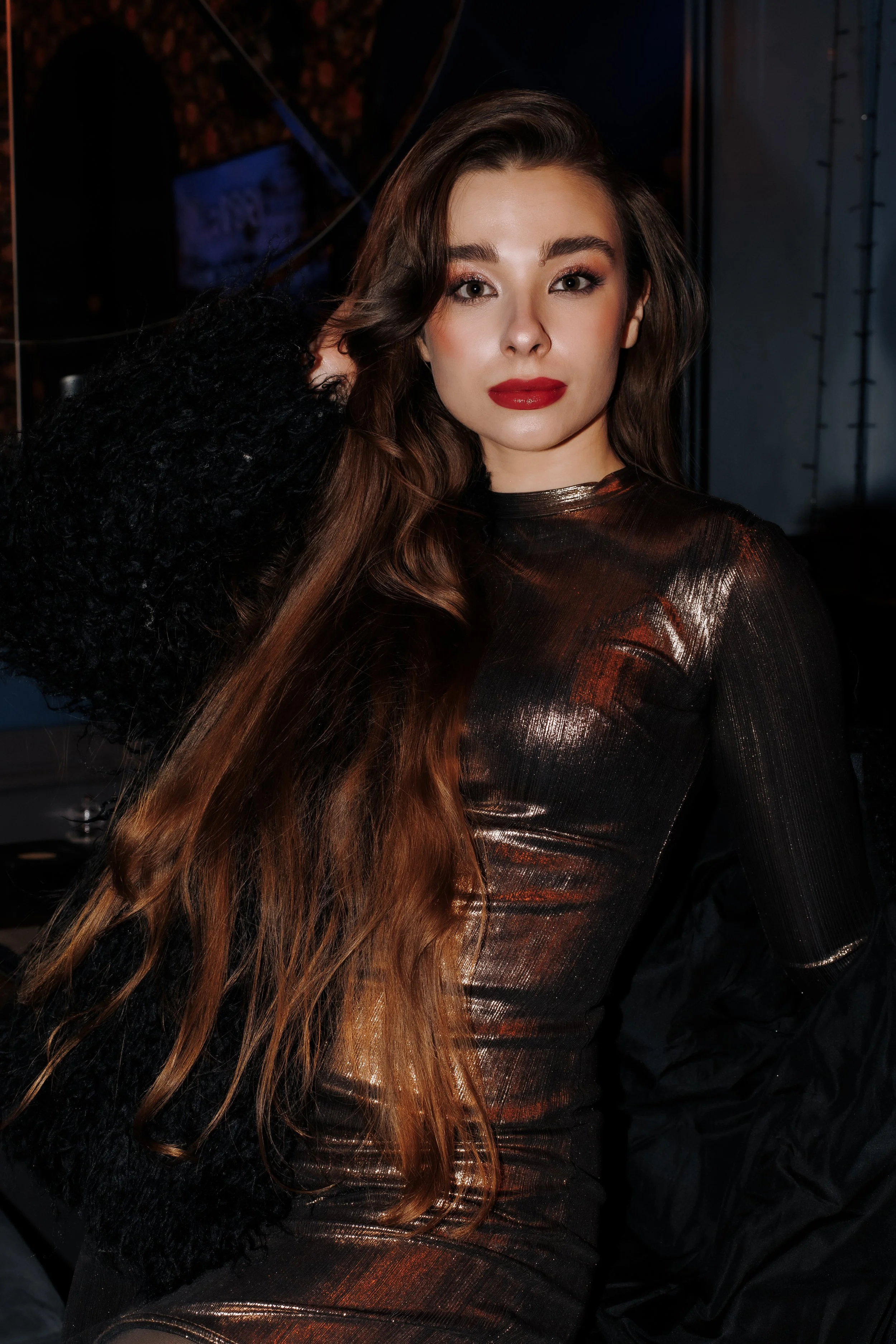 A woman with long wavy brown hair and red lipstick, wearing a shiny black dress, posed in a dimly lit setting.