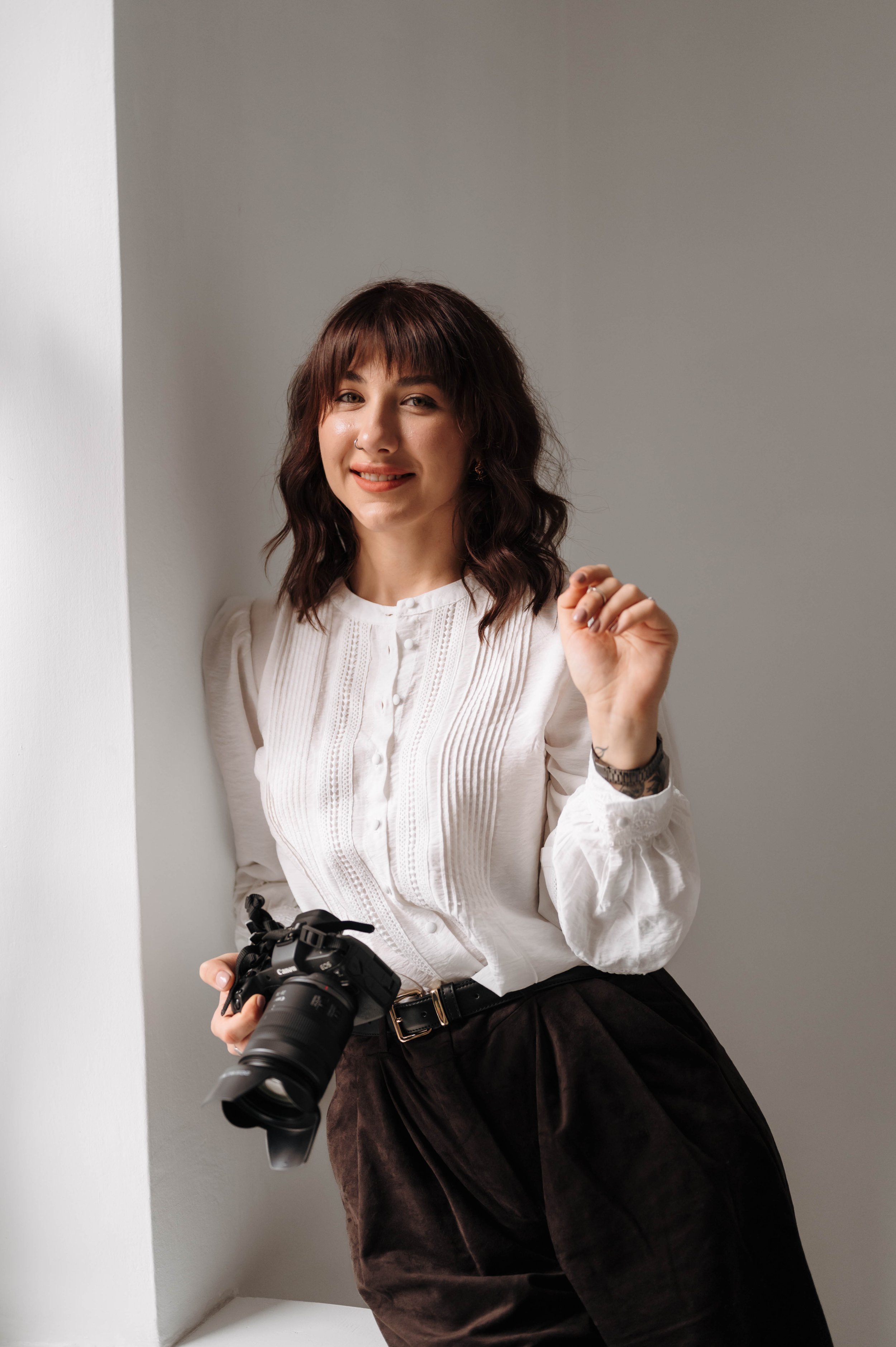A young woman with dark wavy hair and a nose piercing smiling while holding a camera in one hand, standing in a well-lit room near a white wall.