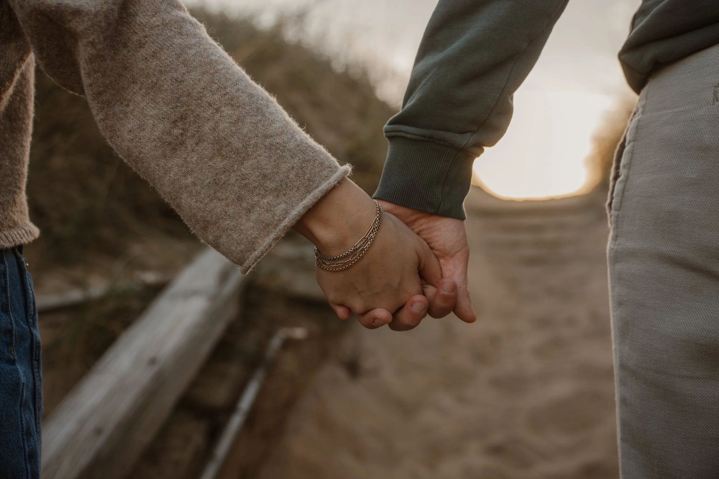 Close-up of two people holding hands, with one wearing a gray sweatshirt and the other wearing a brown jacket, on a dirt path near a wooden fence, with a blurred outdoor background at sunset.