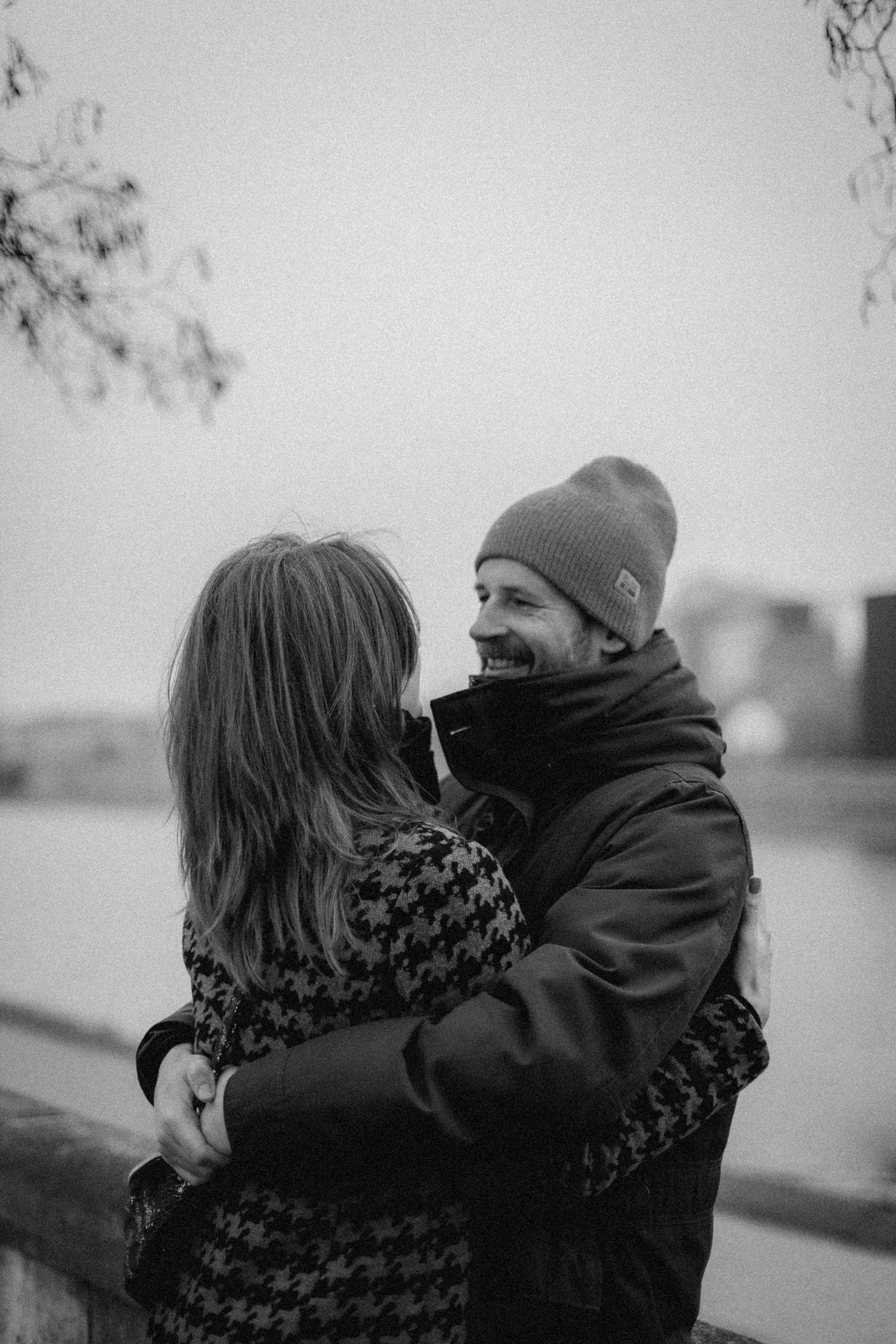 Black and white photo of a couple hugging outdoors near a body of water, with trees in the background. The man is wearing a beanie and a puffy jacket, smiling, while the woman has long hair and a patterned coat.