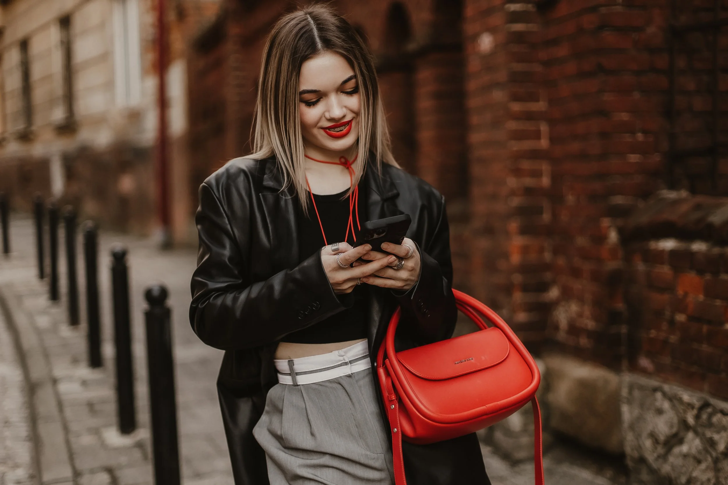Young woman with blonde hair, wearing a black leather jacket, gray trousers, holding a red handbag, smiling at her phone, walking along a brick wall sidewalk.