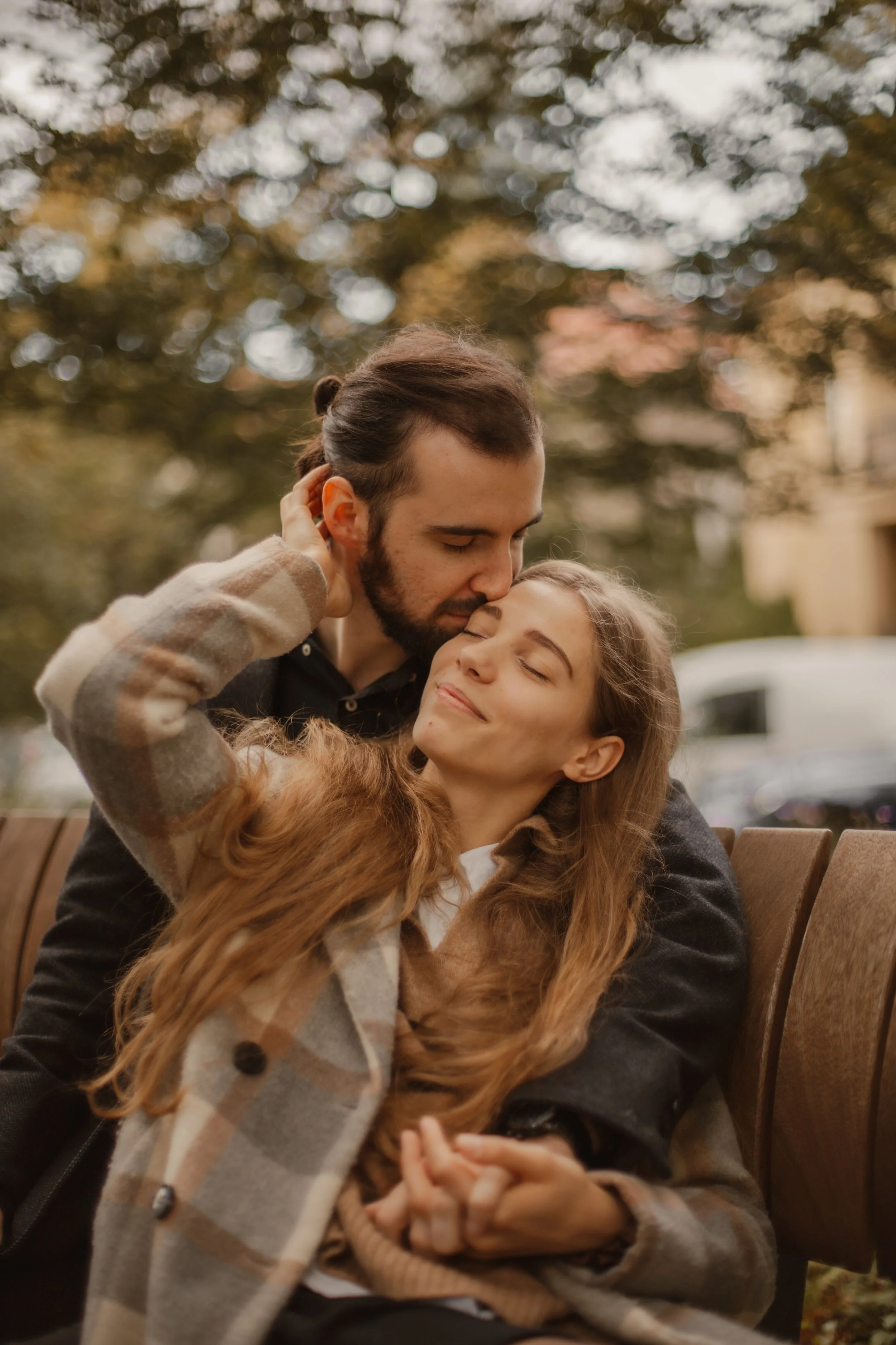 A man and woman sitting on a park bench, sharing a gentle, affectionate moment with eyes closed and smiles, surrounded by a blurred background of autumn trees.