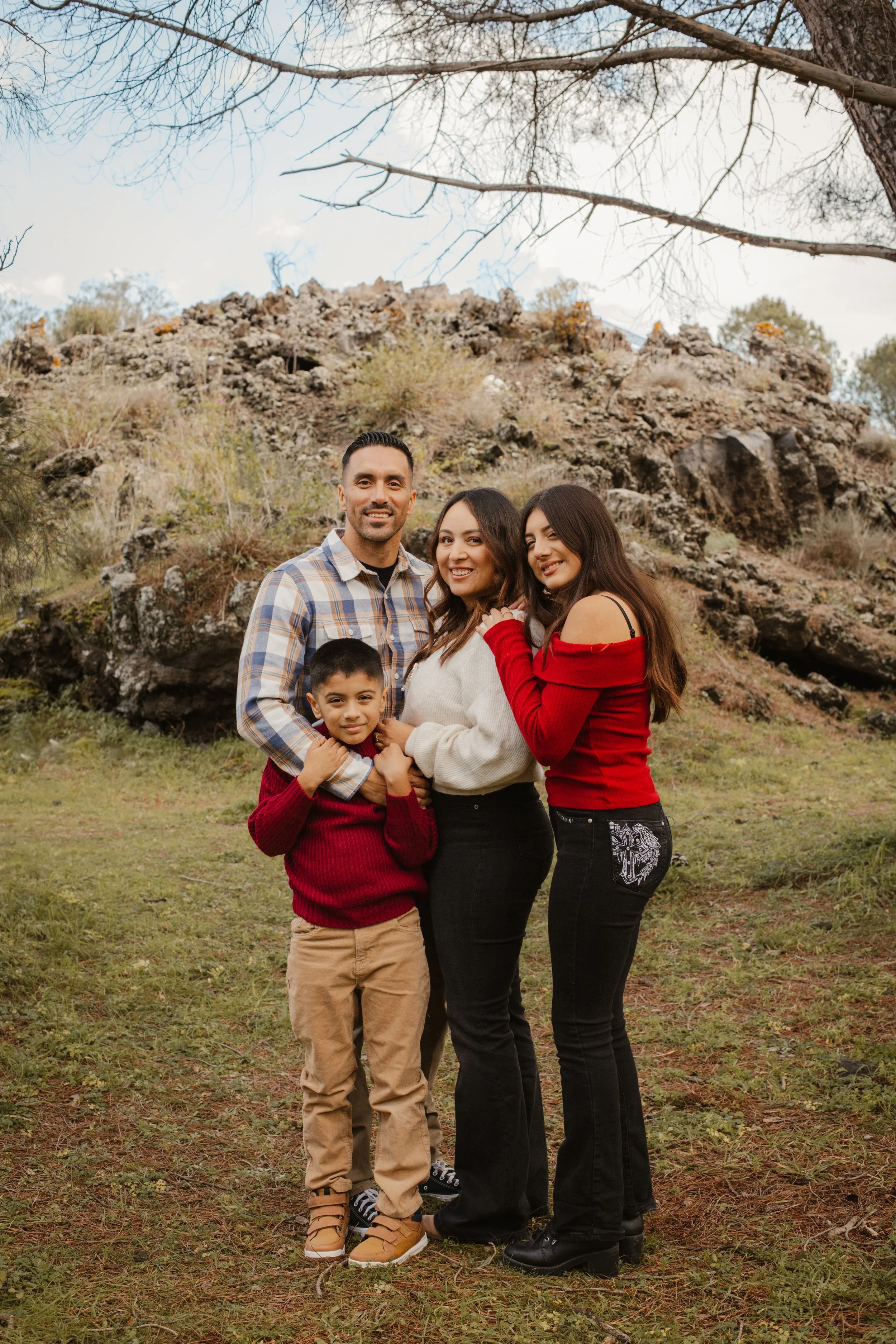 A family of four standing outdoors on grass, posing for a photo. There is a rocky hill in the background and leafless tree branches overhead. The family includes a man, two women, and a young boy, all smiling at the camera.