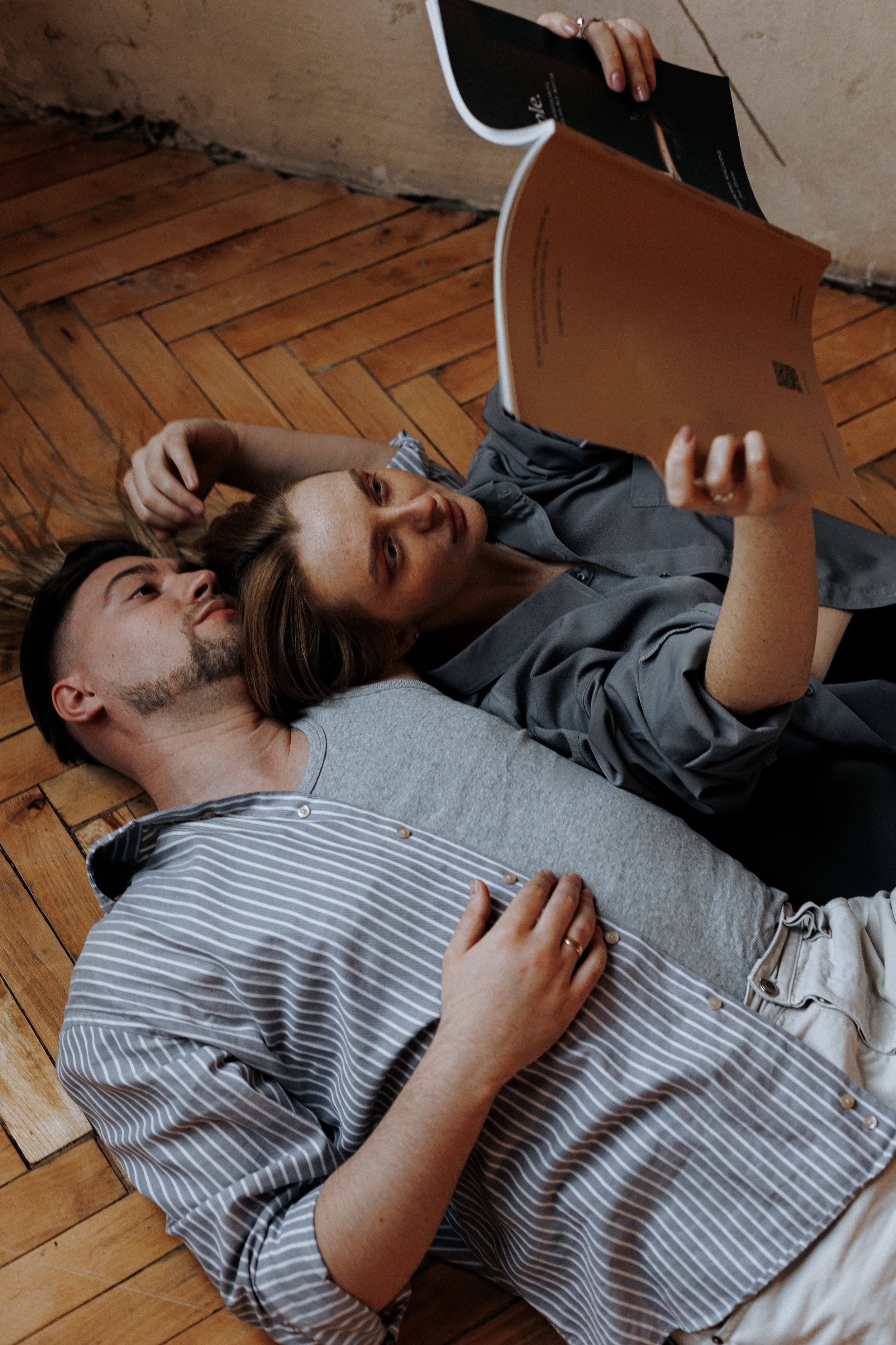 Two people lying on a wooden floor, one reading a magazine and the other resting her head on his shoulder.