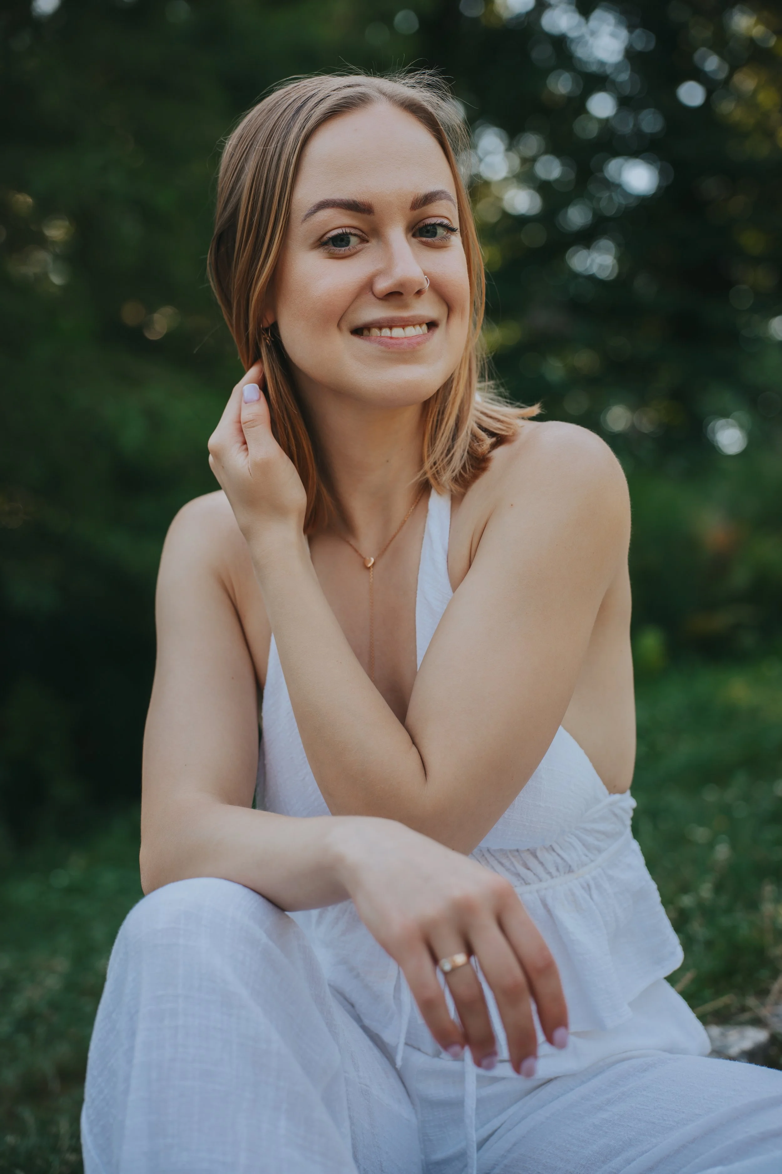 A young woman with shoulder-length light brown hair, smiling and looking at the camera, sitting outdoors in a green setting with blurred trees in the background, wearing a white sleeveless top and matching bottom, with a ring on her left hand and a small necklace.