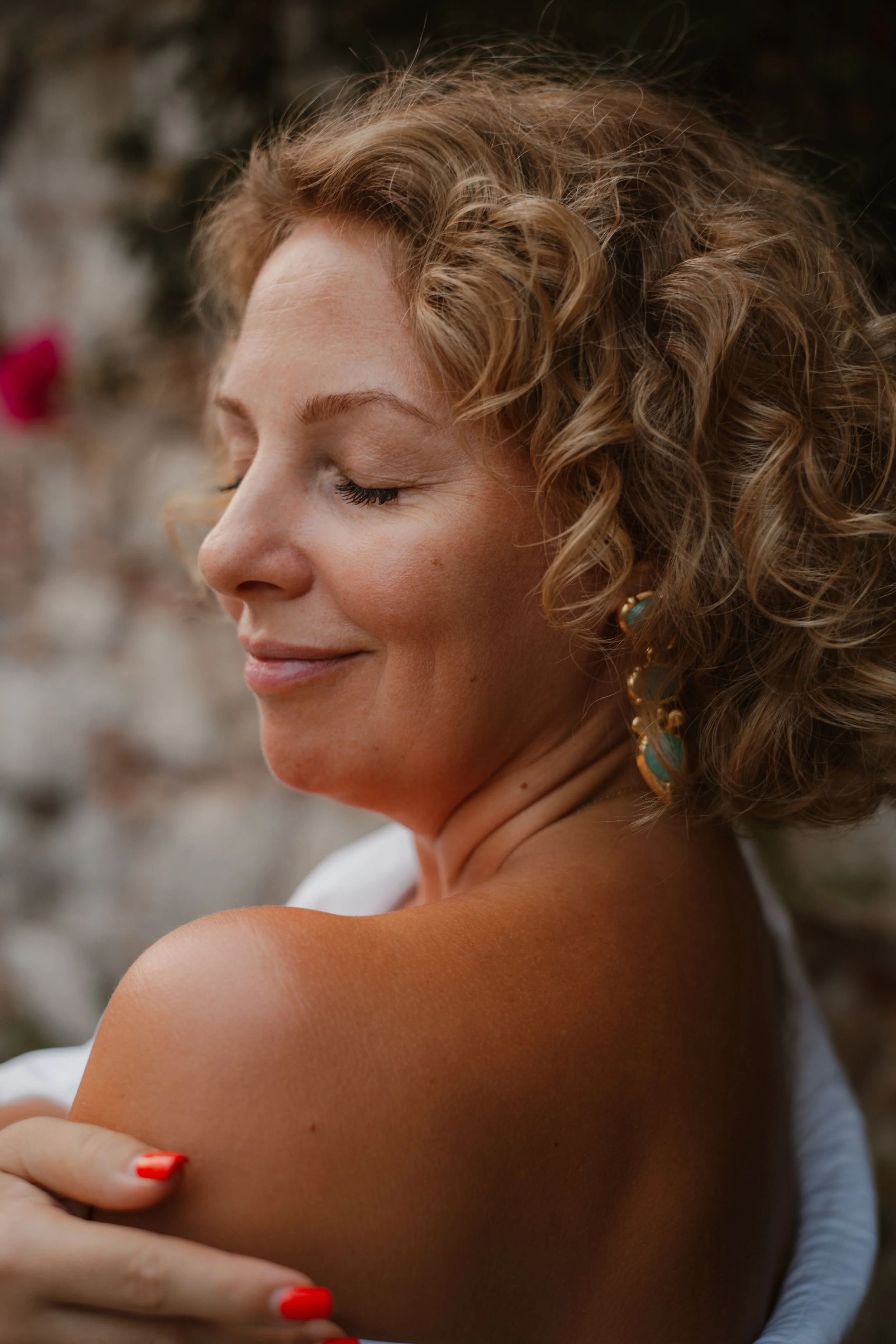 A woman with short, curly blonde hair and earrings, smiling gently with her eyes closed, touching her shoulder.