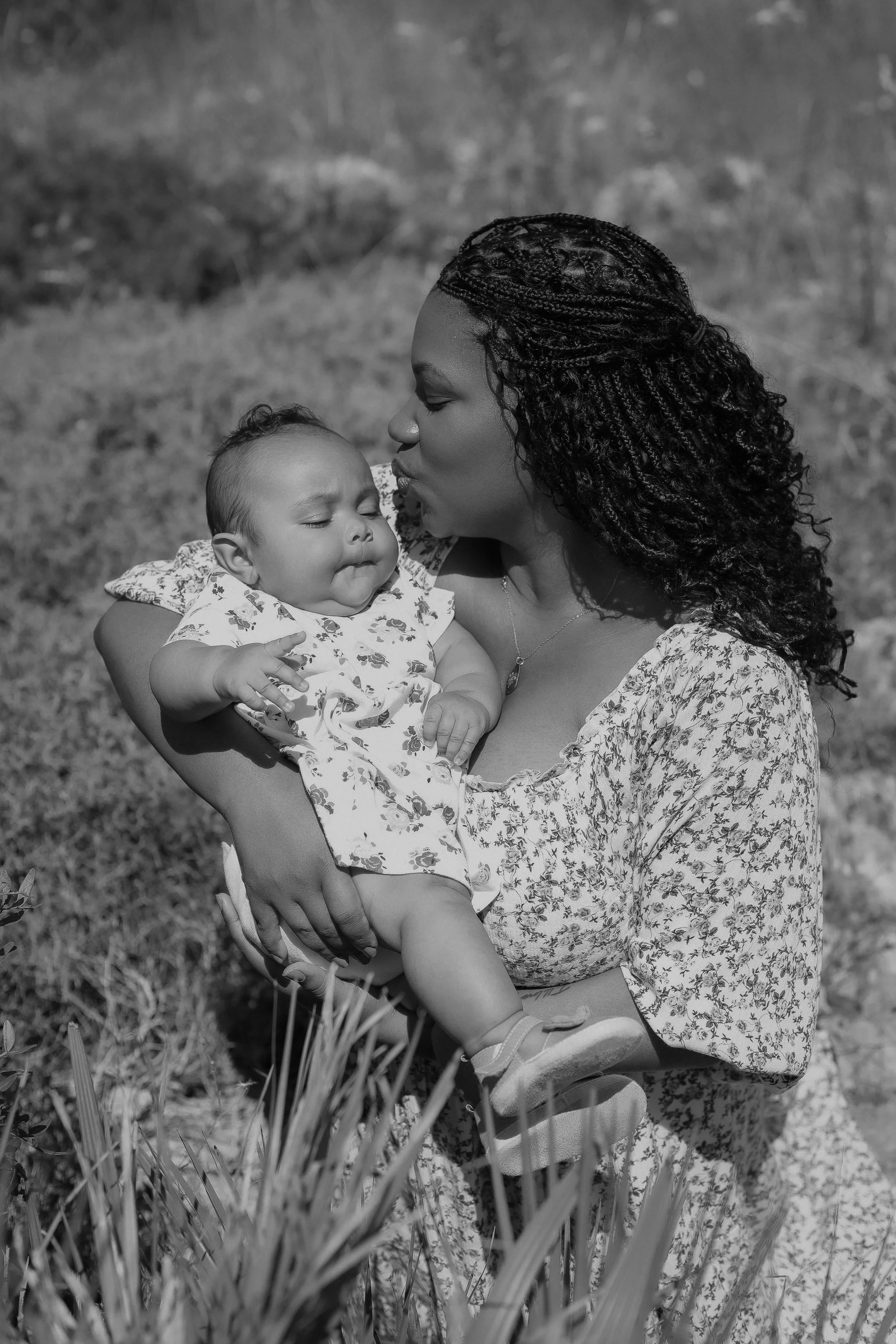 A woman with curly hair holds a baby girl with closed eyes and a content expression in an outdoor setting.