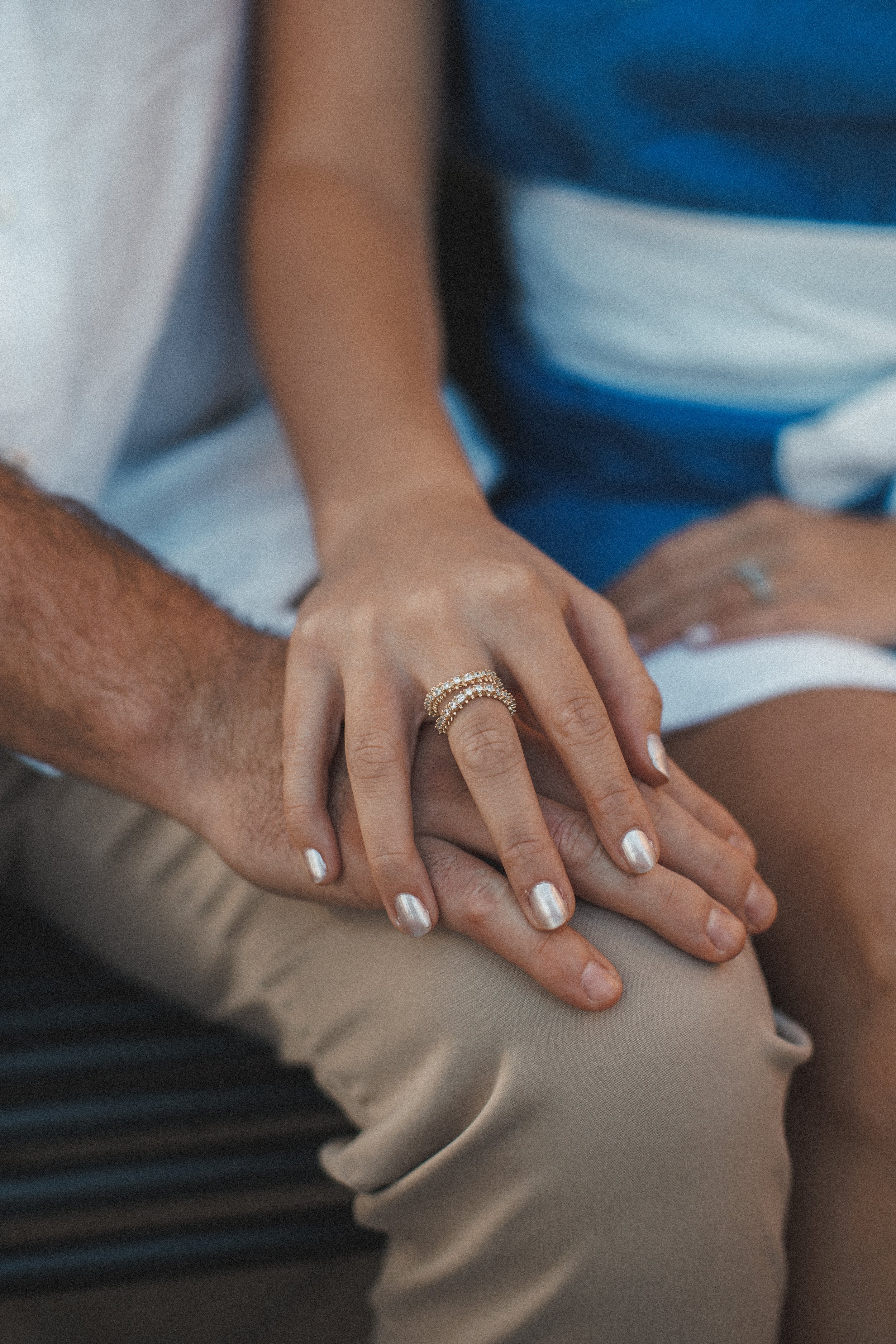 Close-up of a woman's hand with rings, resting on a man's hand, both on a person's lap. The woman has metallic nail polish, and the man has a darker, textured skin tone.
