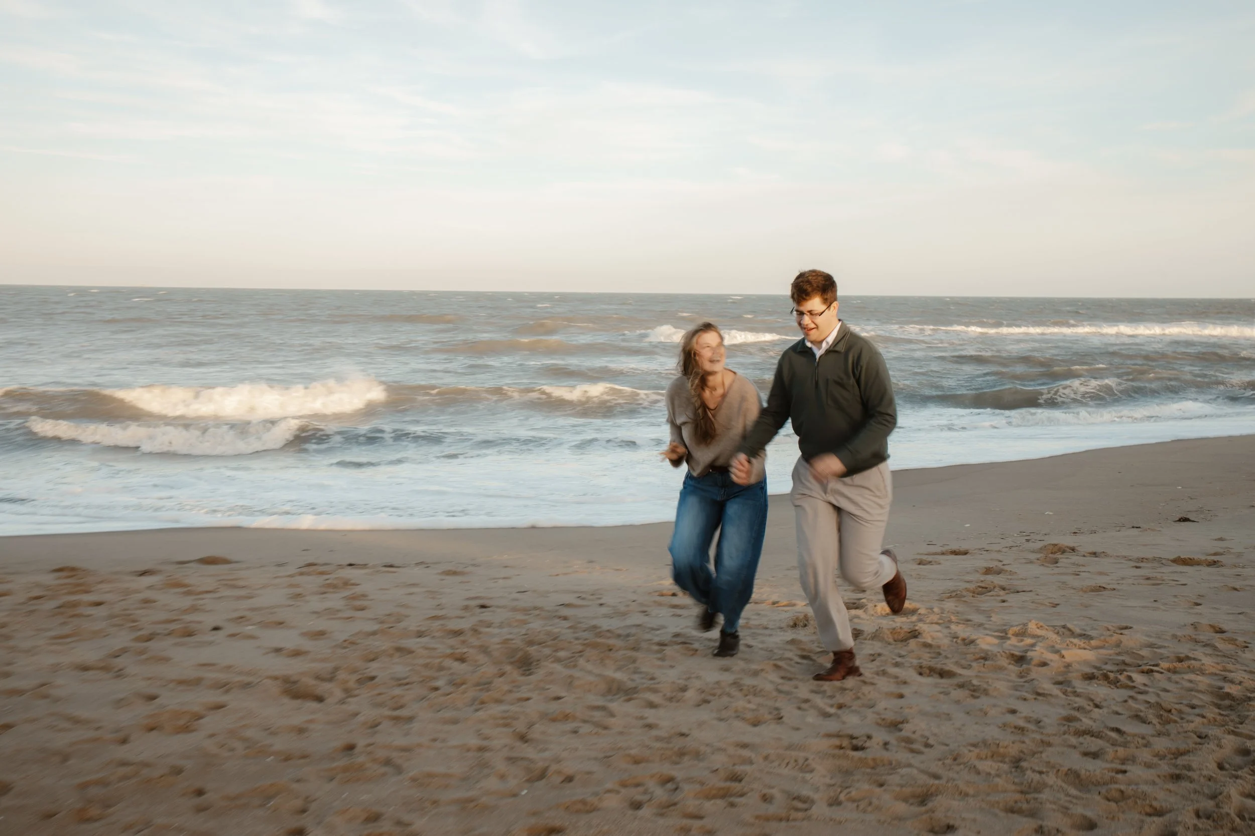 A young man and woman are joyfully running and holding hands on the sandy beach near the ocean waves during the daytime.