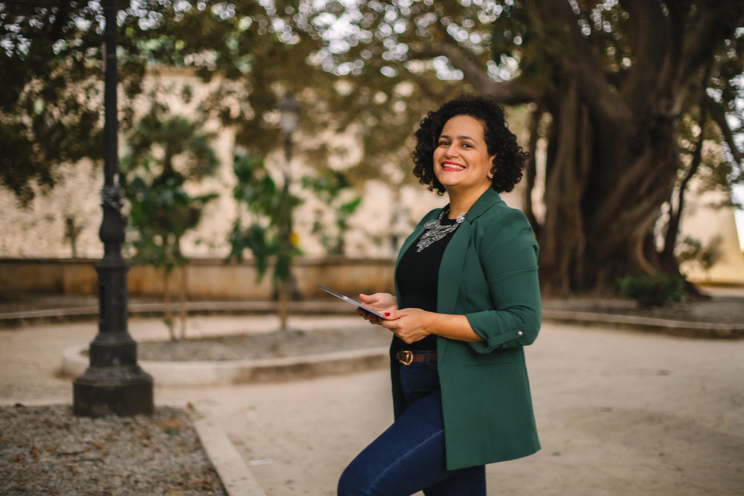 A smiling woman with curly hair wearing a green blazer and dark jeans holding a tablet standing outdoors near a large tree and a vintage-style lamp post.