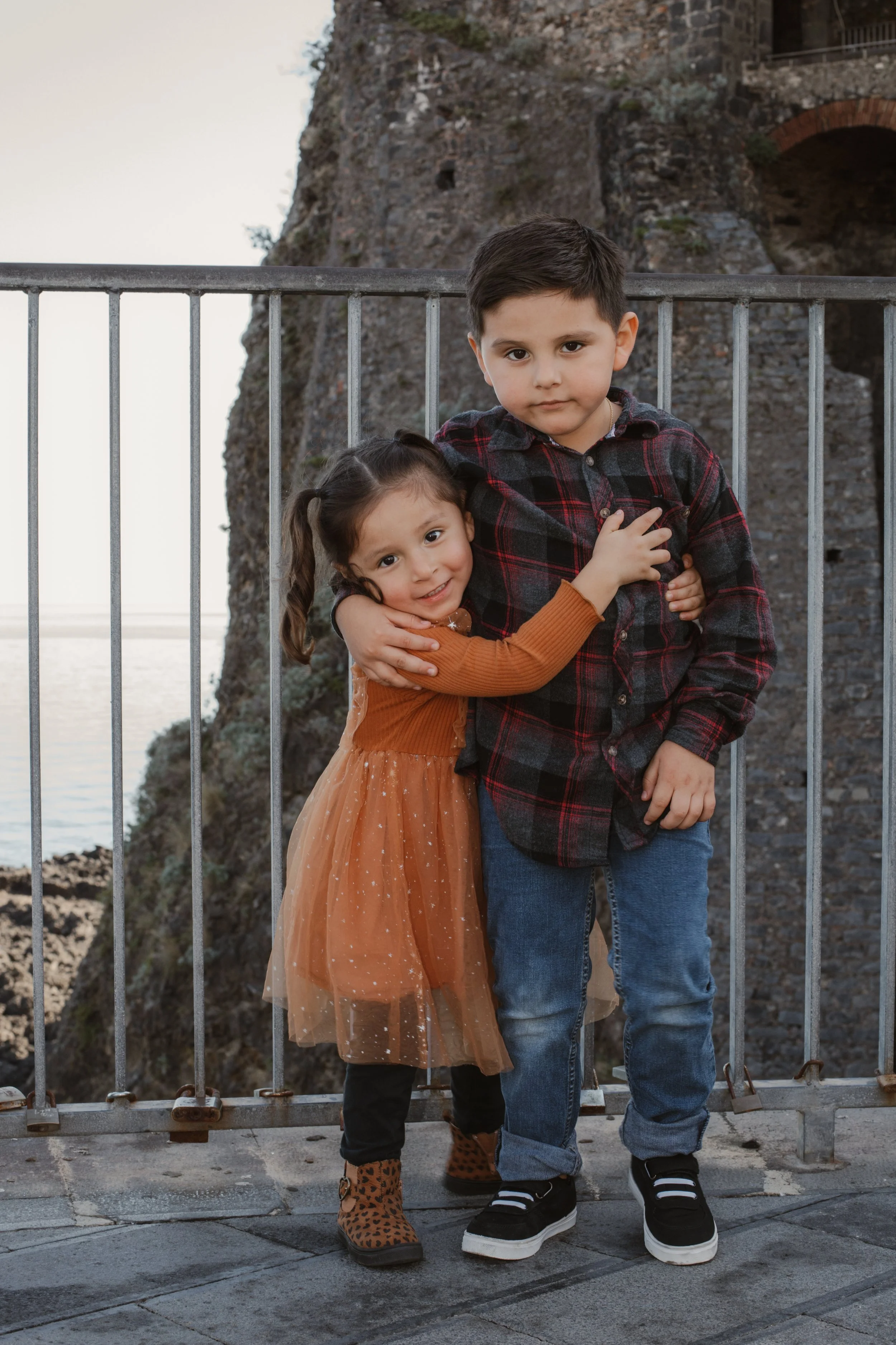 Two children, a girl in an orange dress and leopard print boots hugging a boy in a plaid shirt and jeans, standing by a metal railing with a rocky cliff and ocean in the background.