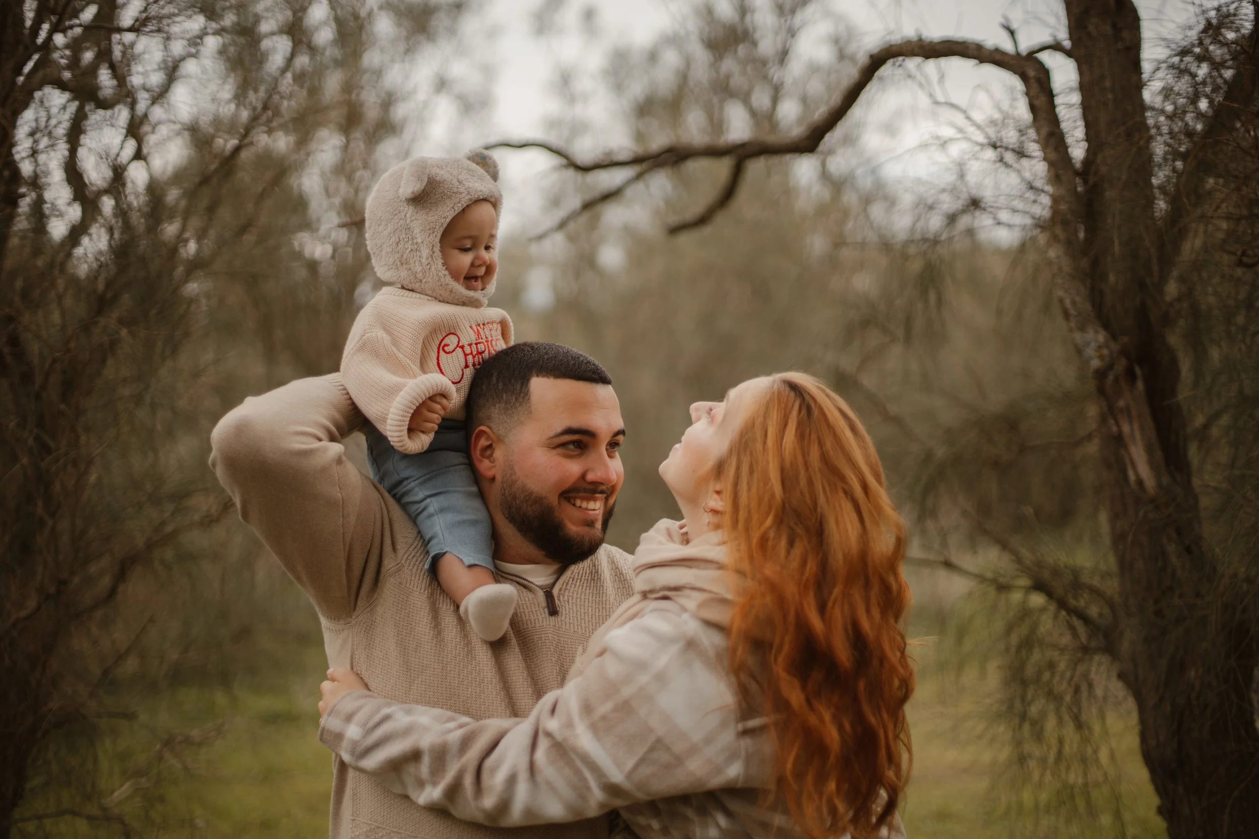 A family of three enjoying outdoors in a wooded area during fall or winter; a man is holding a young girl on his shoulders, and a red-haired woman is reaching up to her, all smiling.