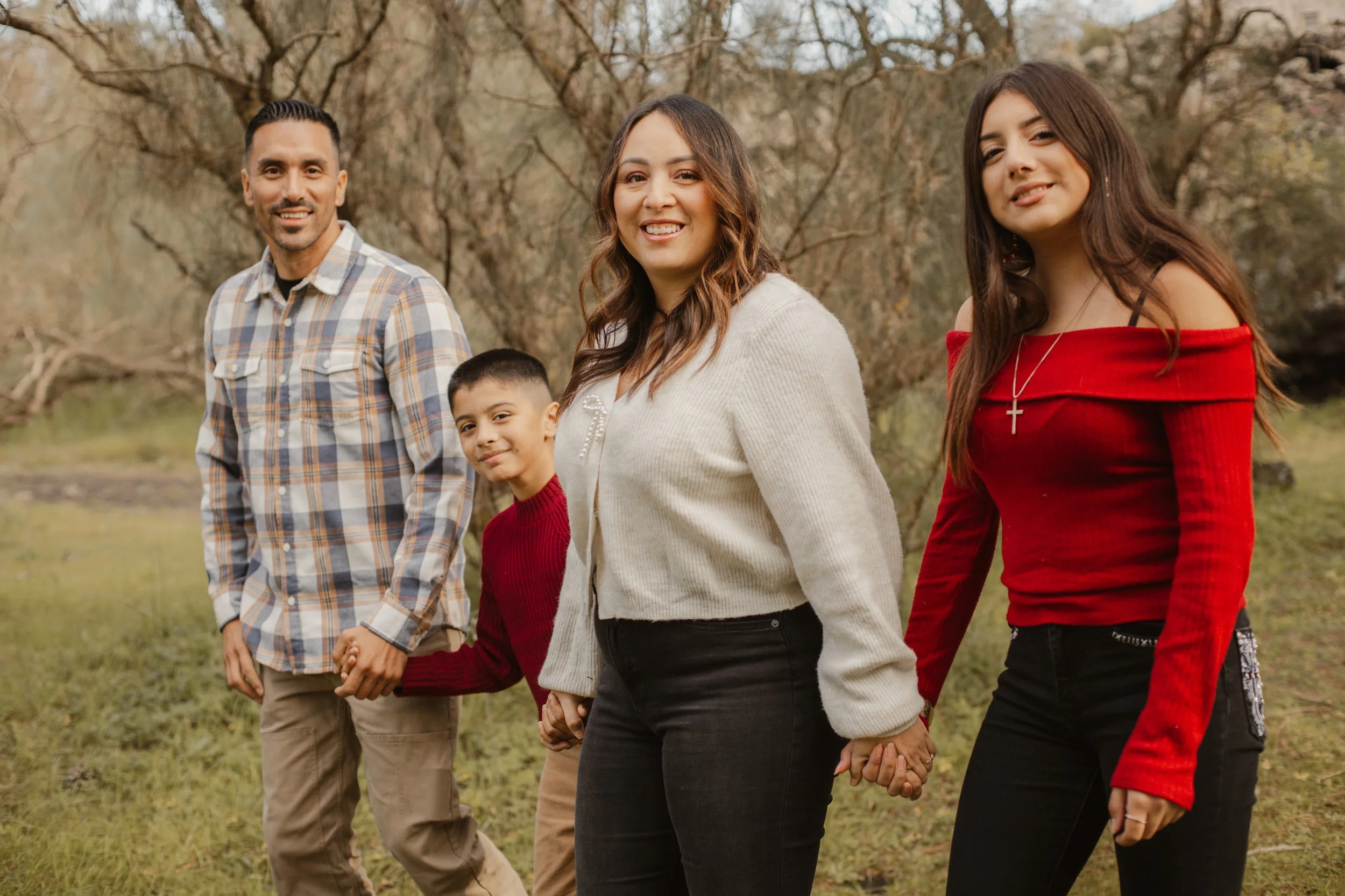 Family of five holding hands outdoors in a park with trees in the background, smiling and looking at the camera.