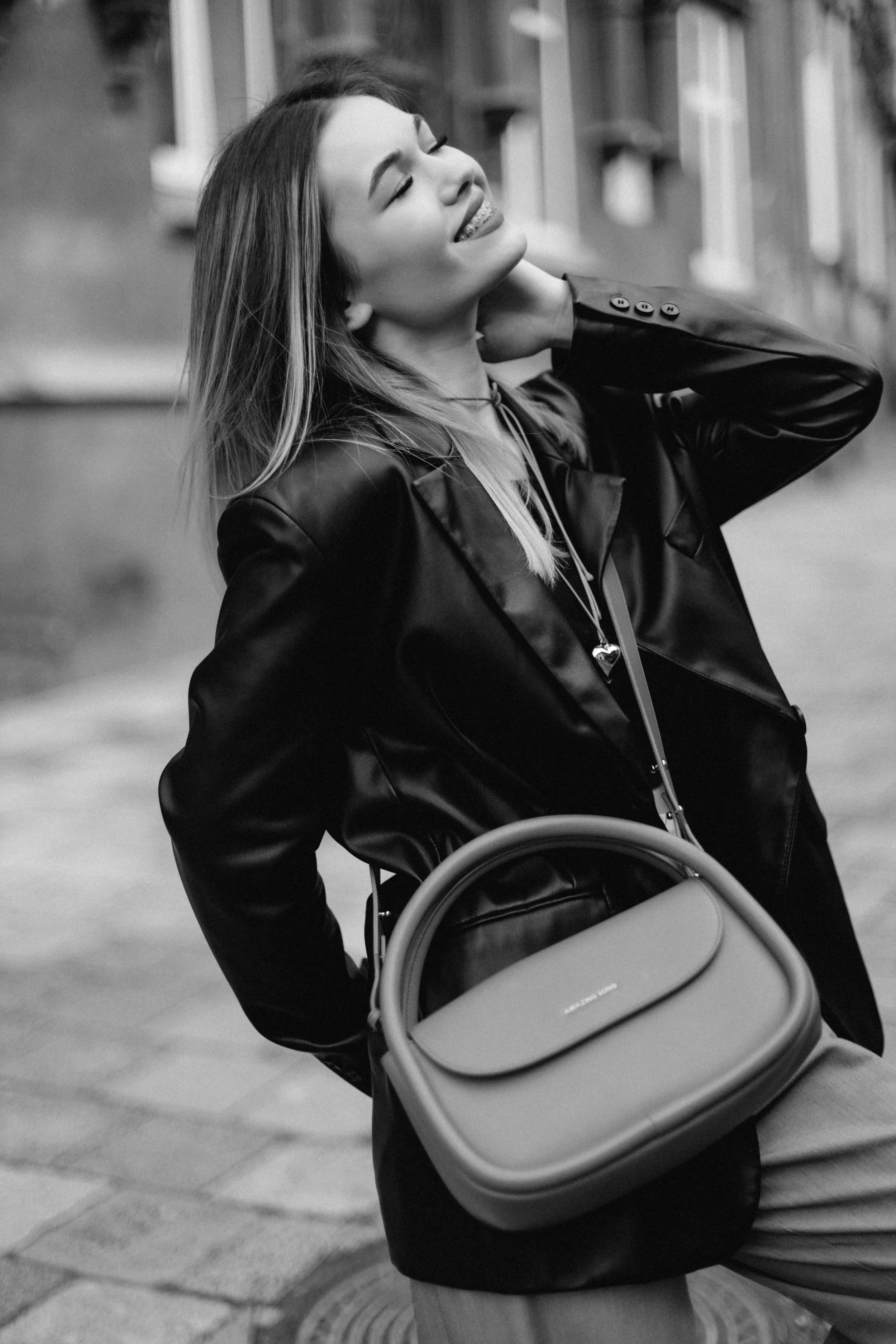 A smiling woman in a leather jacket holding a bag, standing outdoors on a city street.