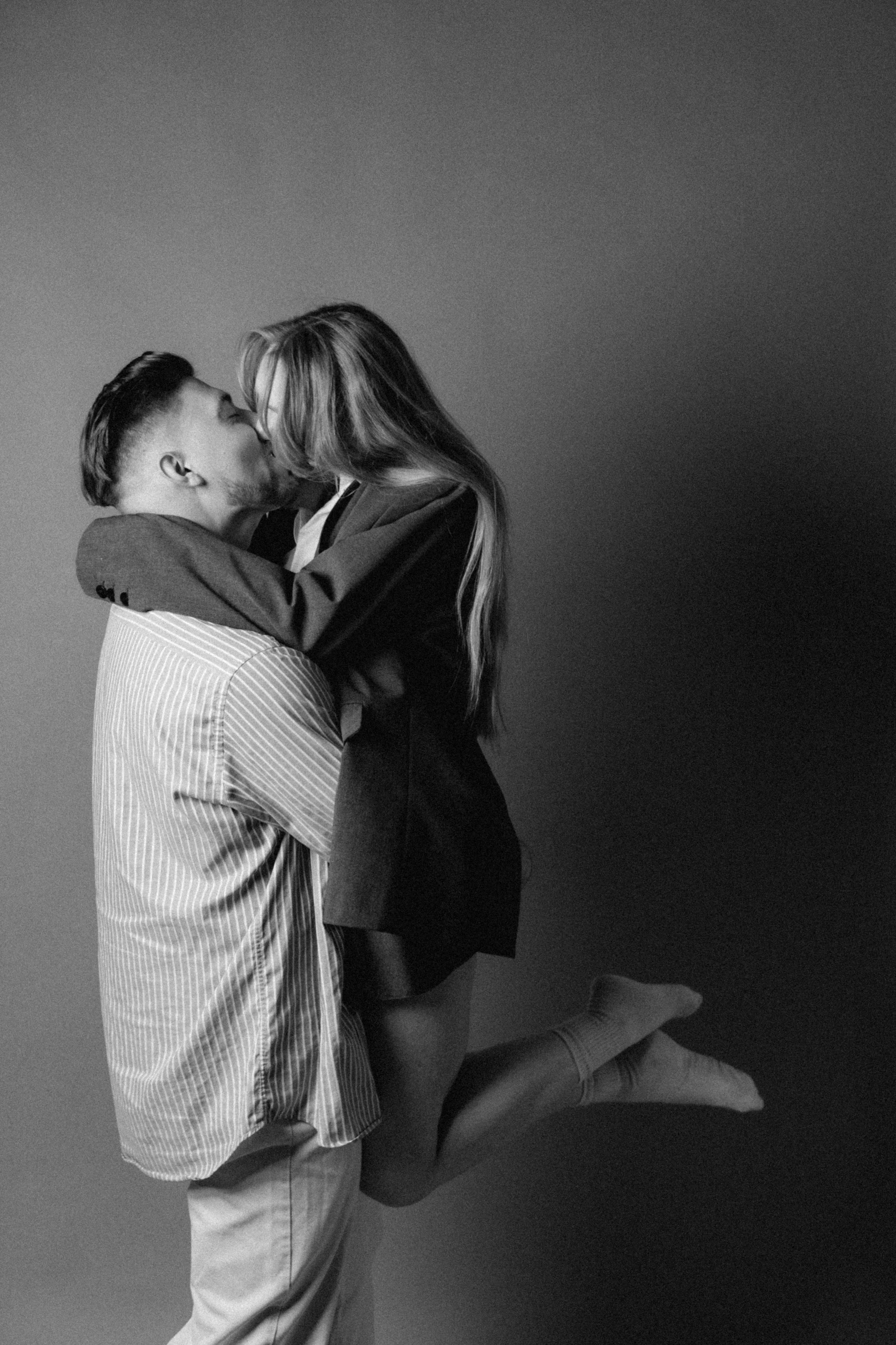 A man lifting a woman in his arms, they are kissing against a plain background, black and white photography.