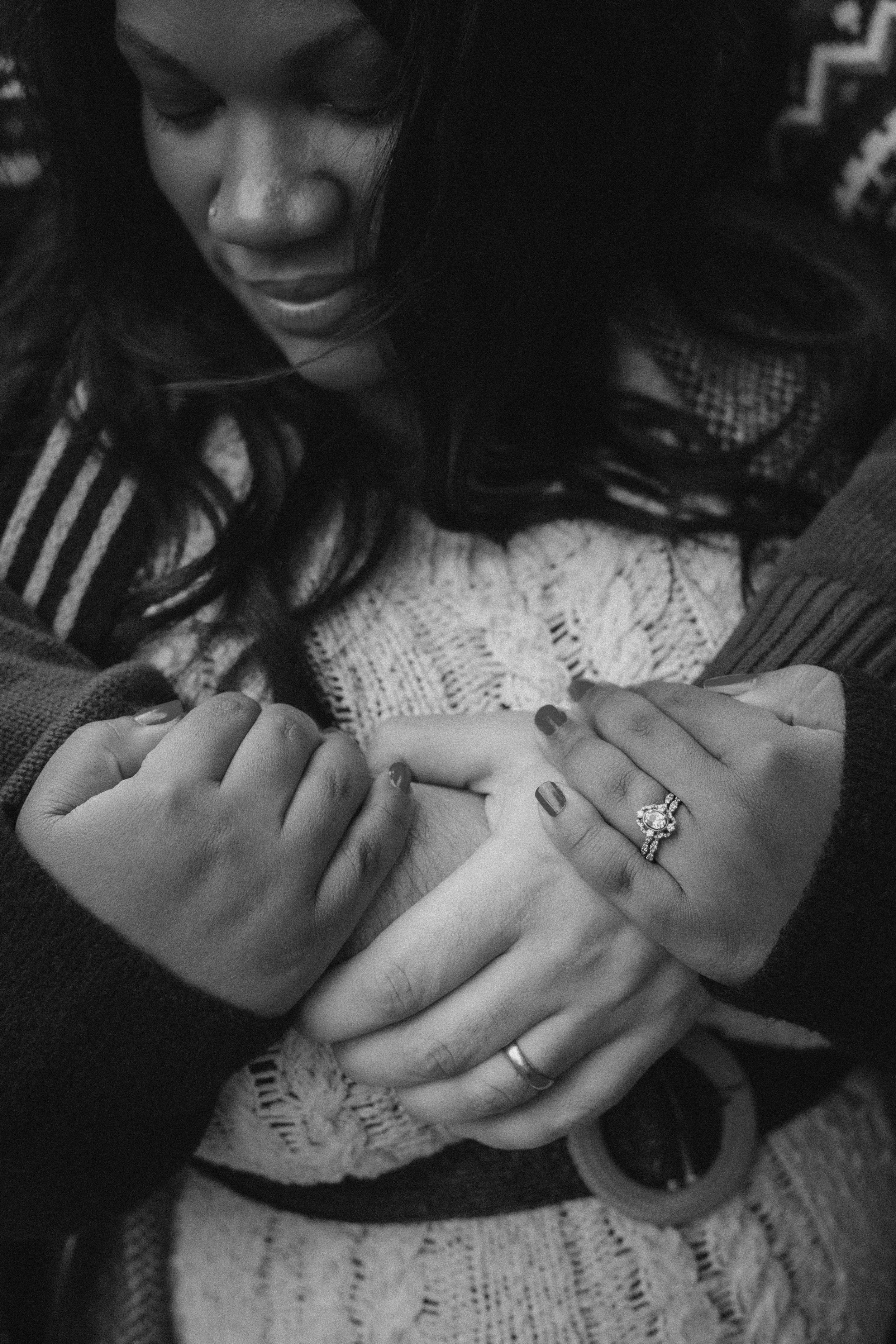 A woman with dark hair and a nose piercing is looking down, holding hands with a person showing a wedding ring on their finger, in black and white.