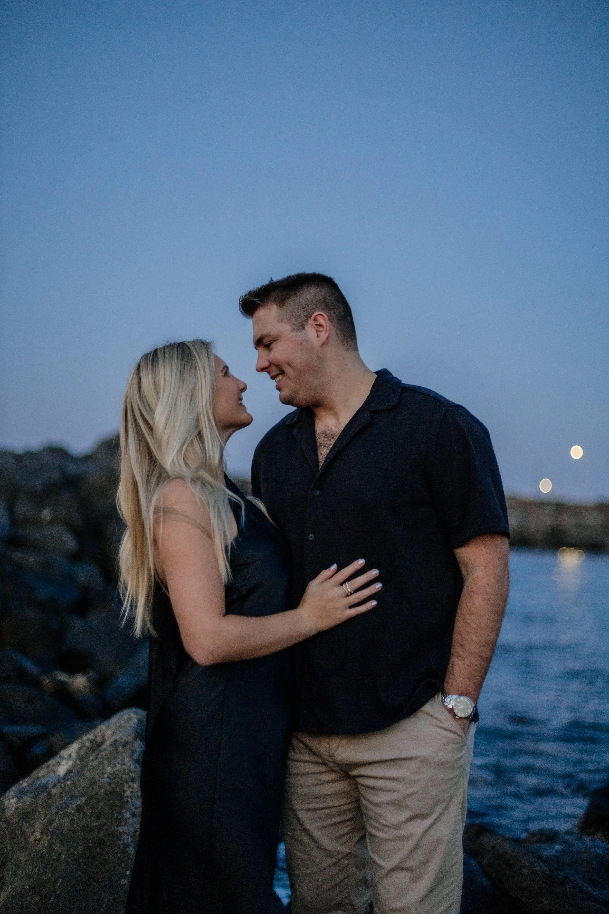 A couple standing close together by the water during twilight, smiling and gazing into each other's eyes.