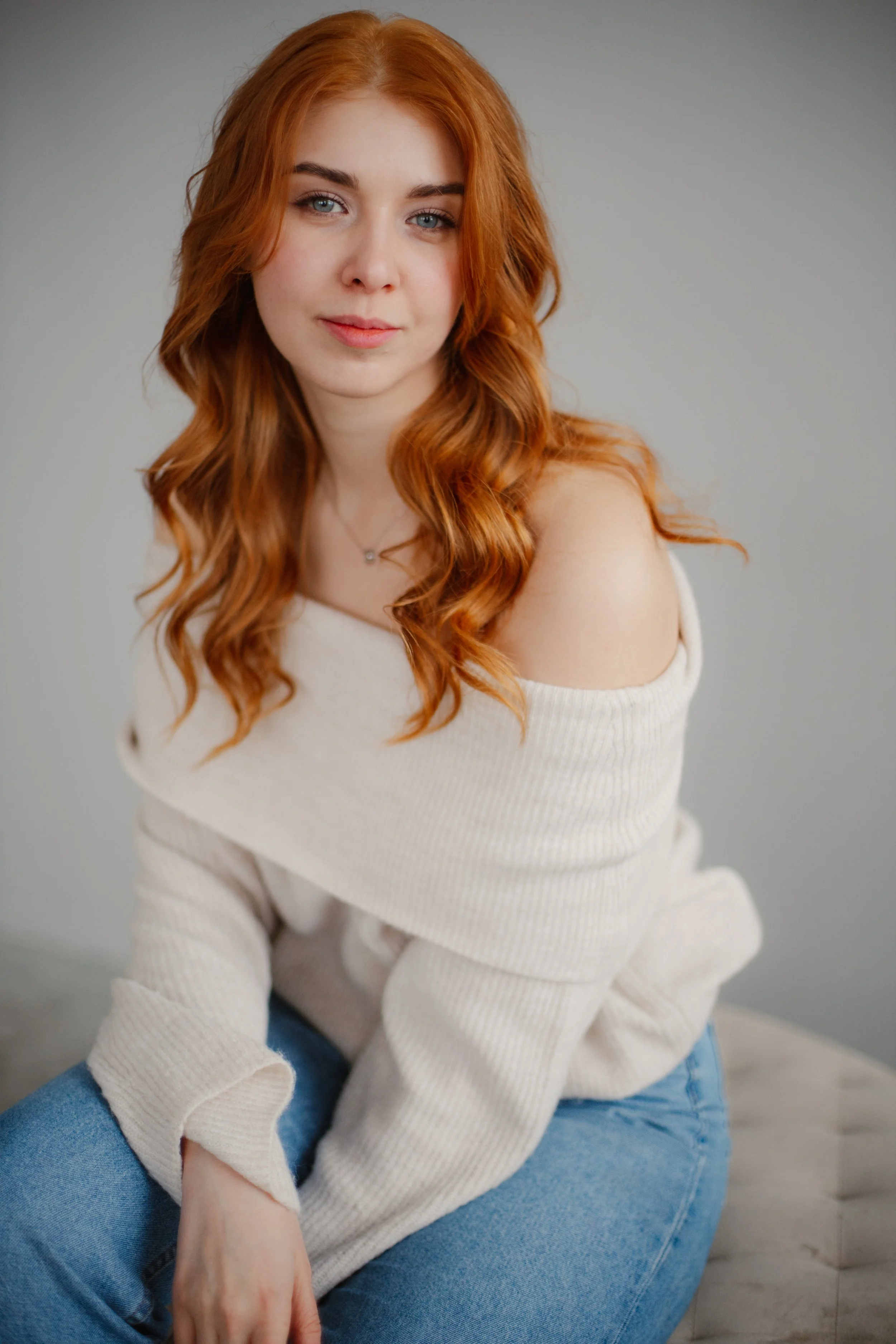A young woman with long, wavy red hair and blue eyes sitting indoors against a plain gray background, wearing a cream off-the-shoulder sweater and blue jeans.