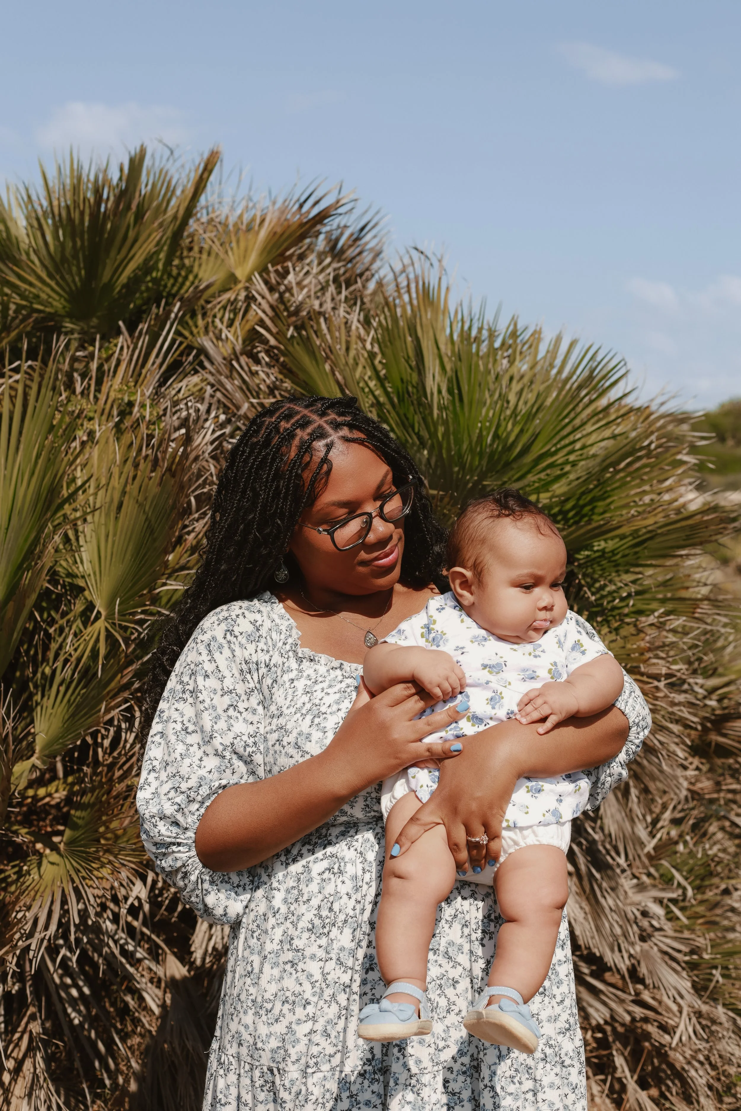 A woman holding a young child outdoors with palm trees and blue sky in the background.