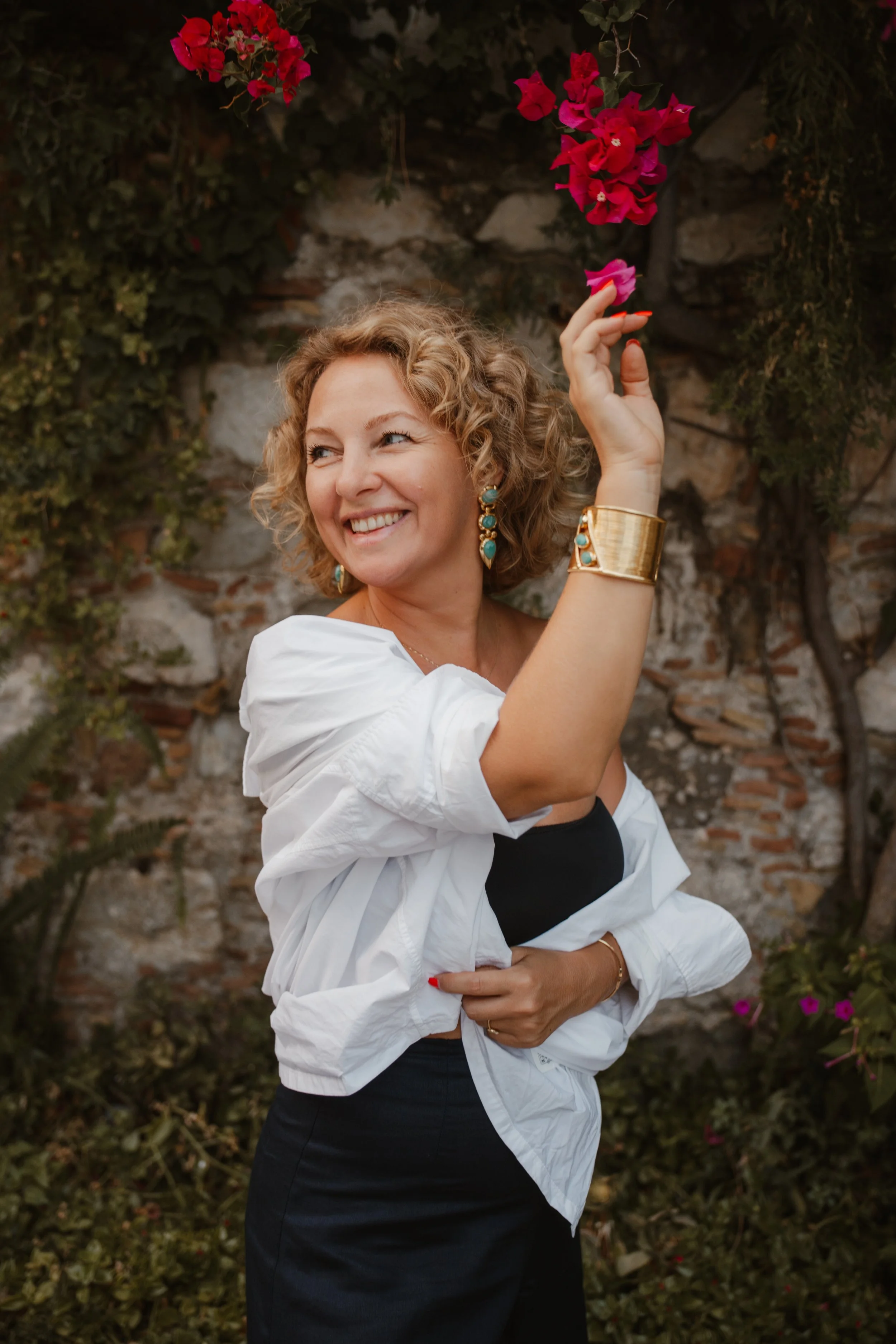 A woman with curly blonde hair, wearing a black top, white jacket, and gold jewelry, smiling and holding a pink flower, standing outdoors in front of a brick and stone wall with flowers.