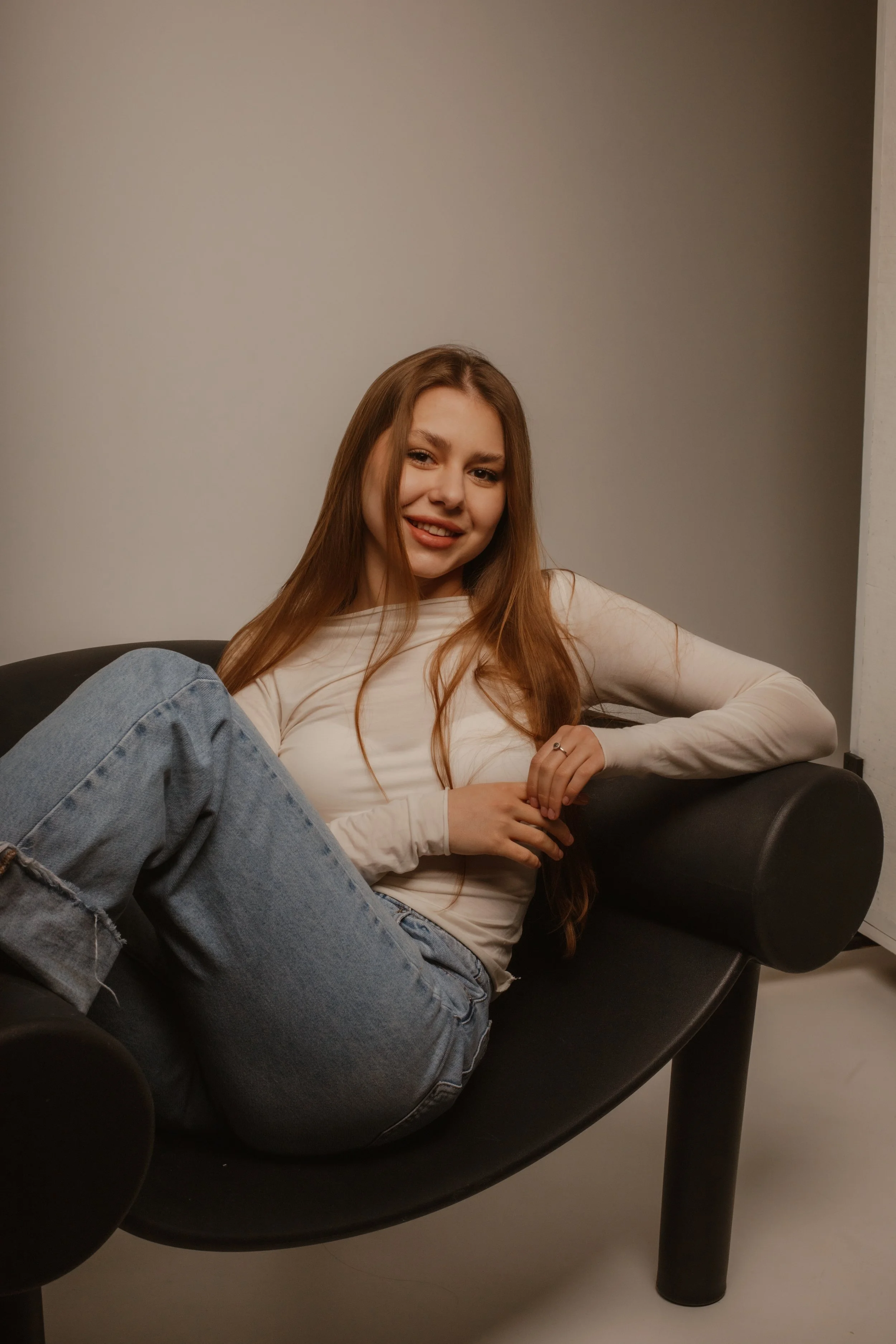 A young woman with long red hair sitting on a black sofa, smiling slightly, wearing a white long-sleeve shirt and light blue jeans.