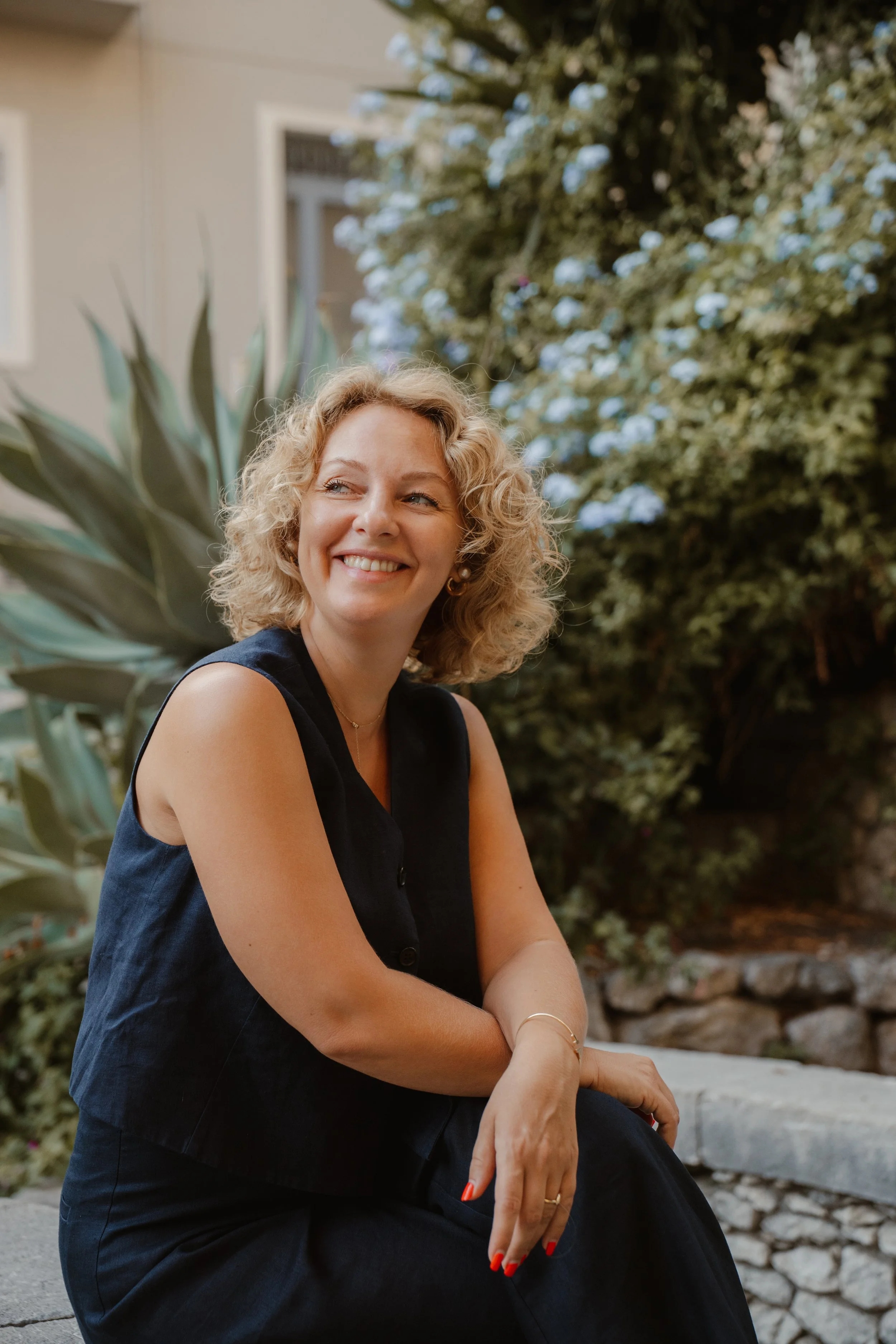 A woman with blonde curly hair smiling outdoors surrounded by greenery and plants.