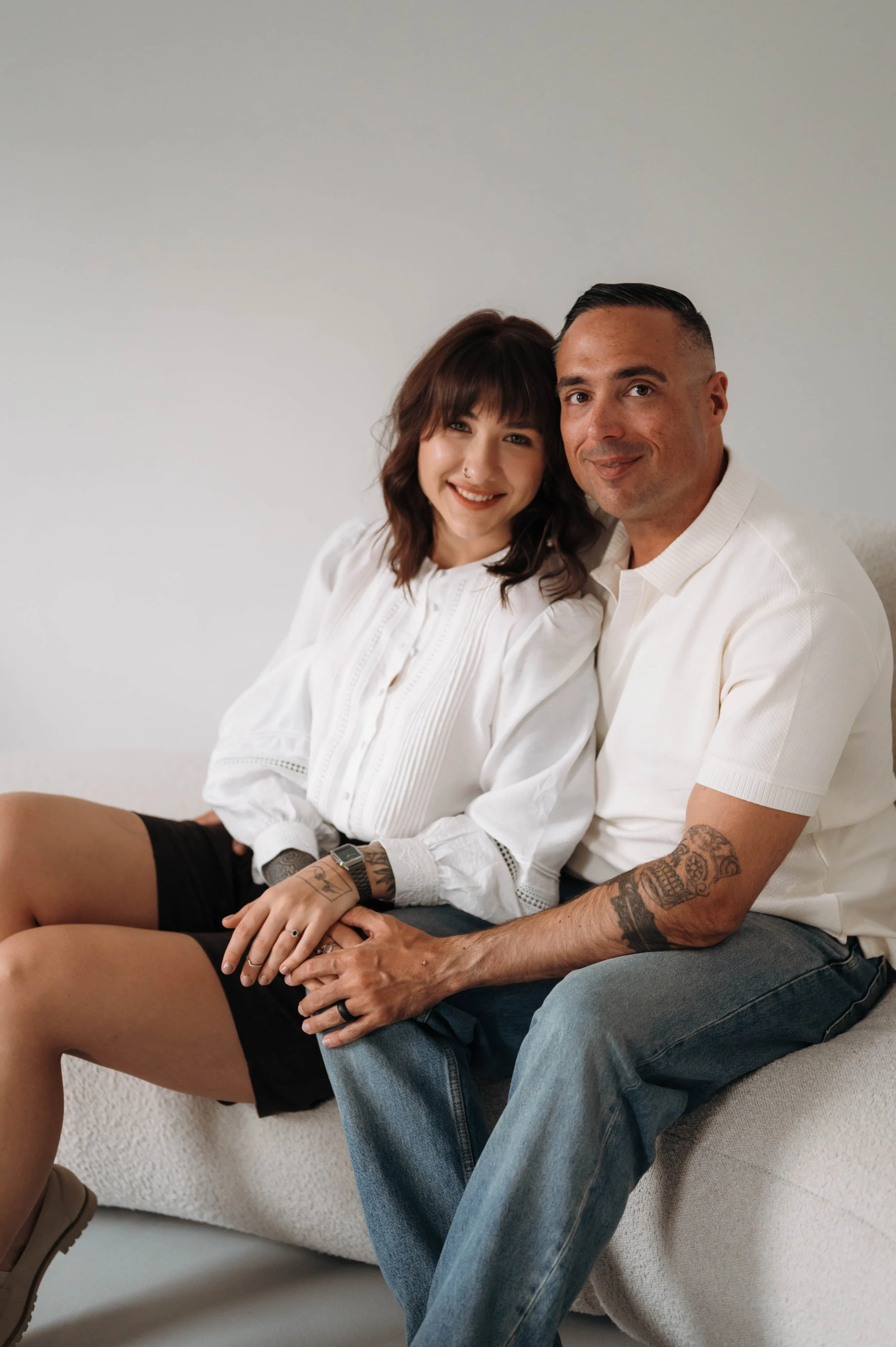 A smiling young woman with brown hair and tattoos sitting with a man with tattoos, both wearing white shirts, on a white sofa against a plain white wall.
