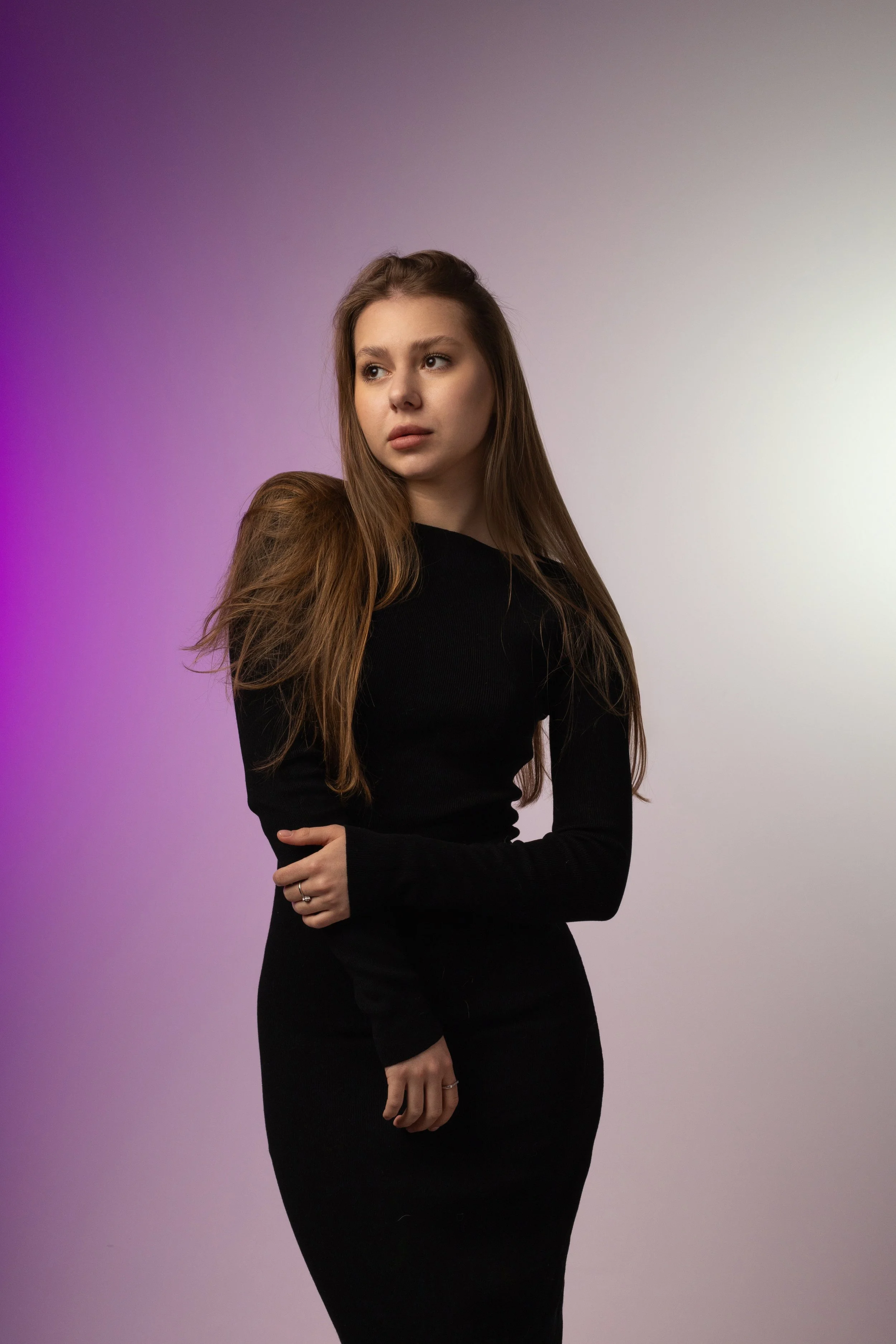 A young woman with long brown hair wearing a black long-sleeve top and skirt, posing with her arms crossed against a gradient background of purple and light yellow.
