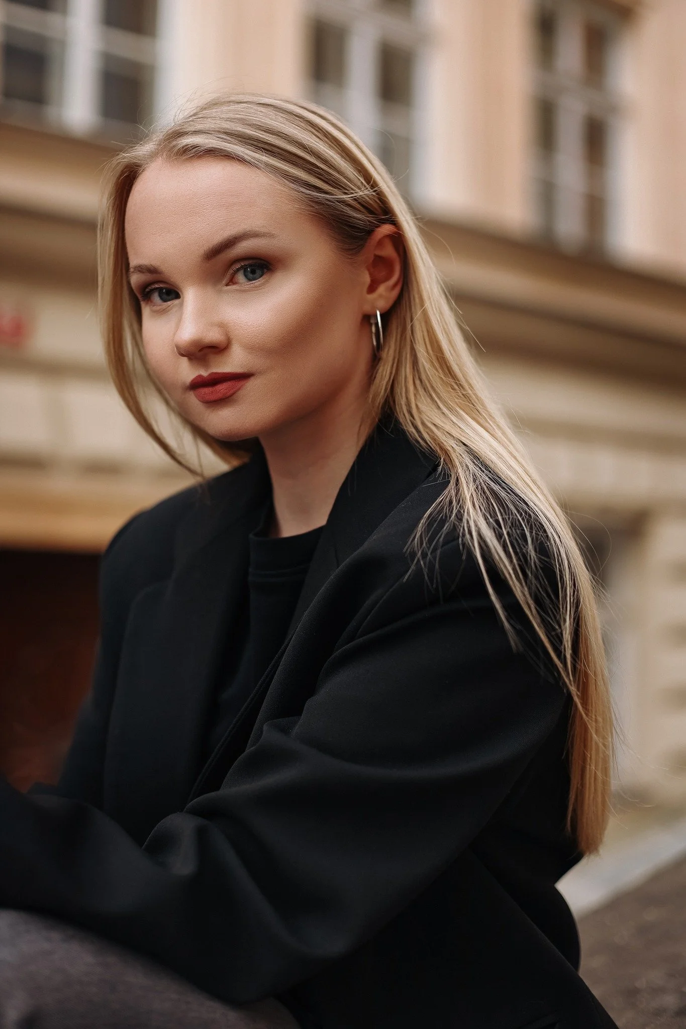 A young woman with blonde hair and blue eyes wearing a black blazer, sitting outdoors with a neutral expression, in an urban setting with a beige building in the background.