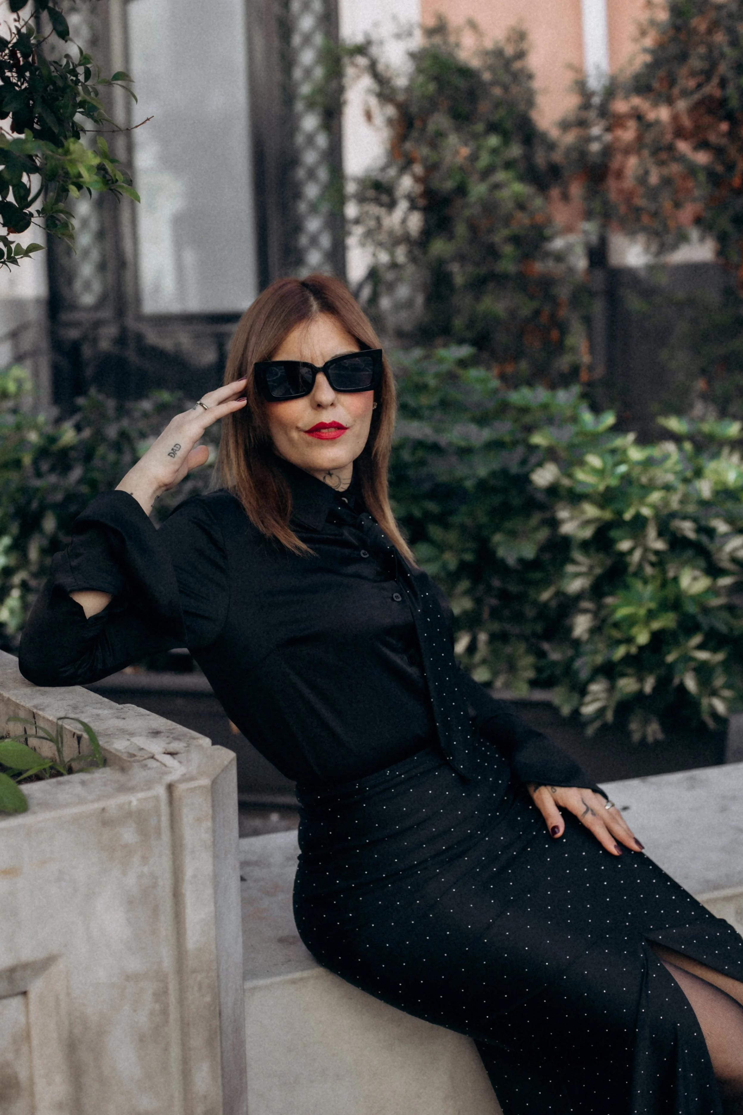 A woman with shoulder-length brown hair wearing black sunglasses, a black shirt, and a black skirt with white polka dots, sitting outdoors on a concrete ledge surrounded by green bushes and trees.