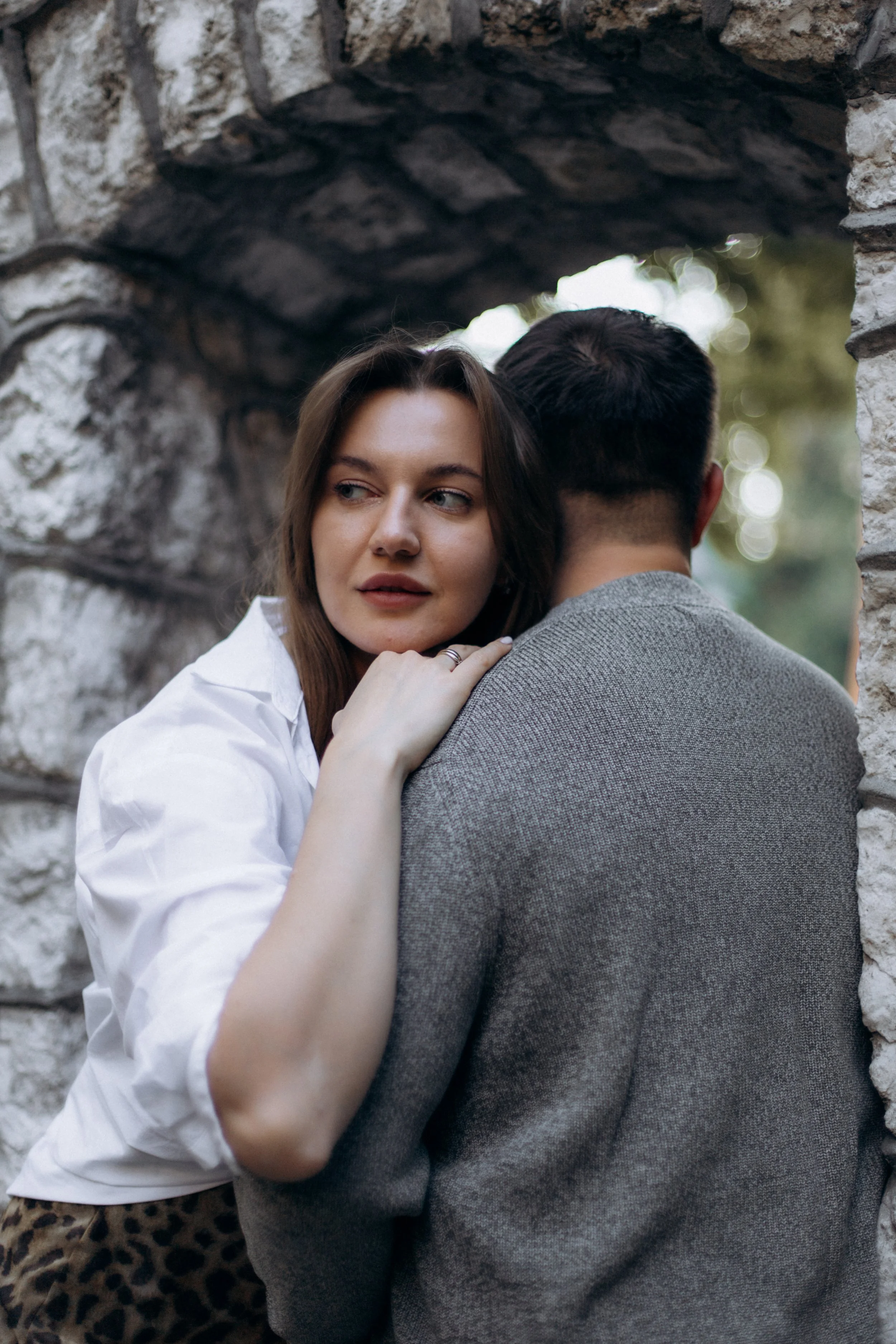 A woman with long brown hair, wearing a white shirt, leaning on the shoulder of a man with dark hair, in a gray textured sweater, near a stone wall opening, outdoors with blurred greenery in the background.
