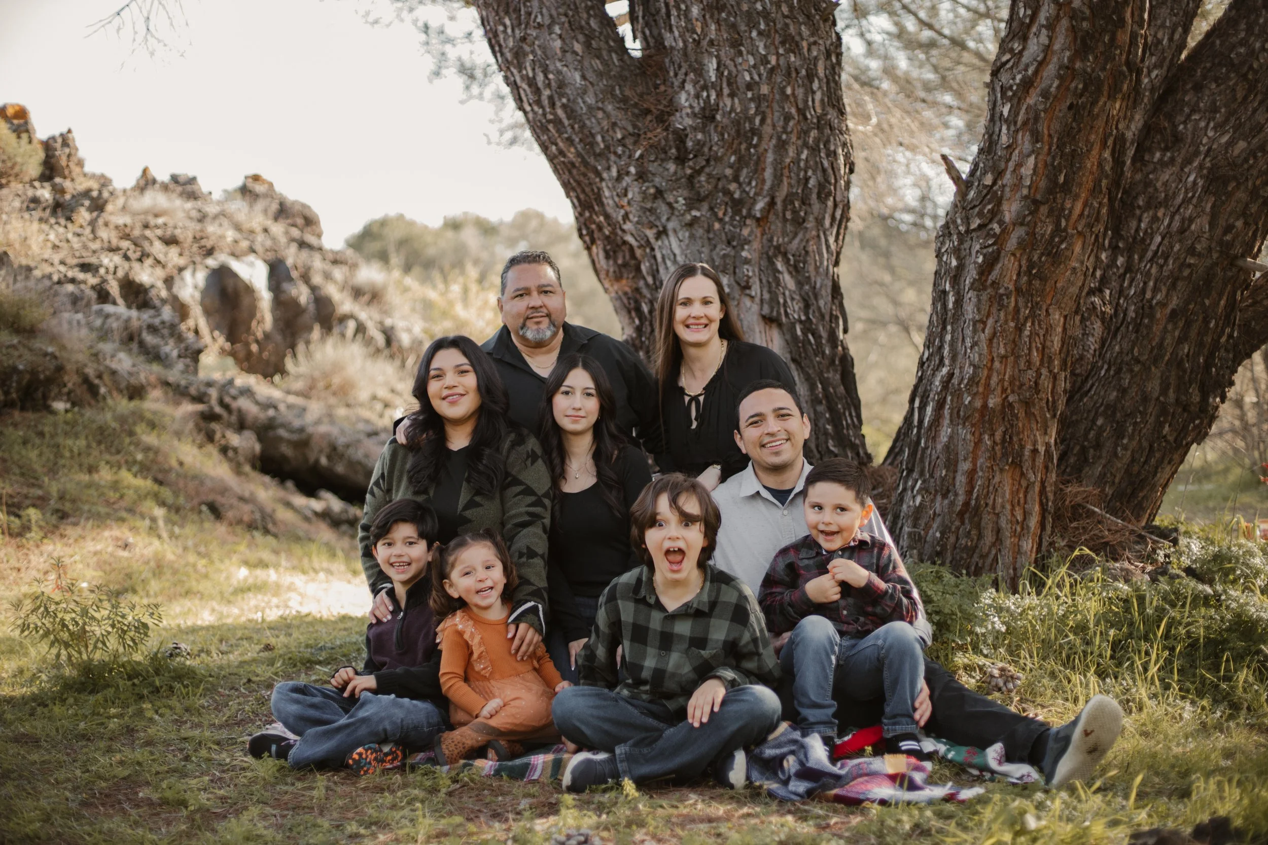 A large family posing outdoors in front of a tree, with children sitting and kneeling on a blanket in front and adults standing behind them.