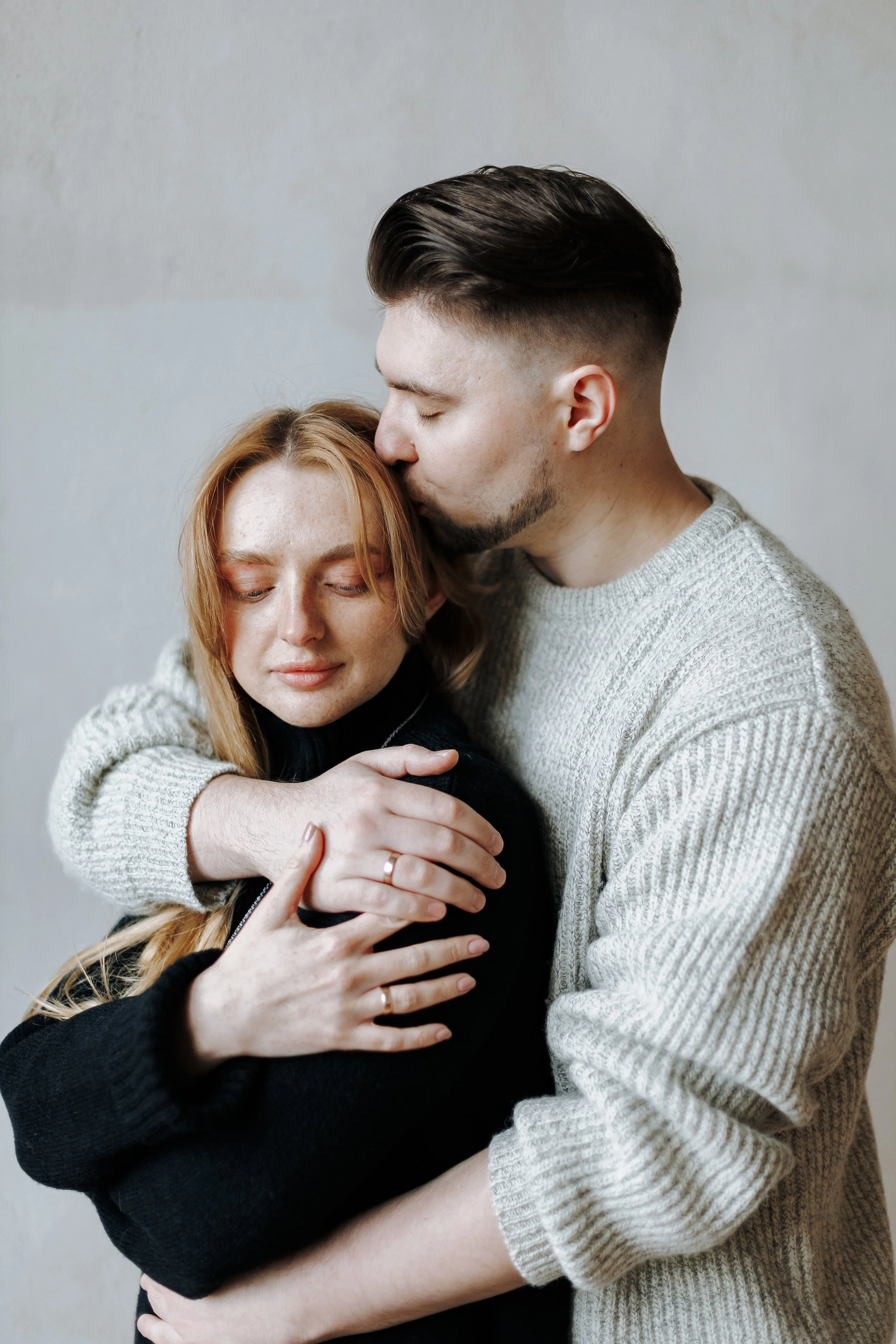 A couple hugging, with the man kissing the woman's forehead against a plain wall.