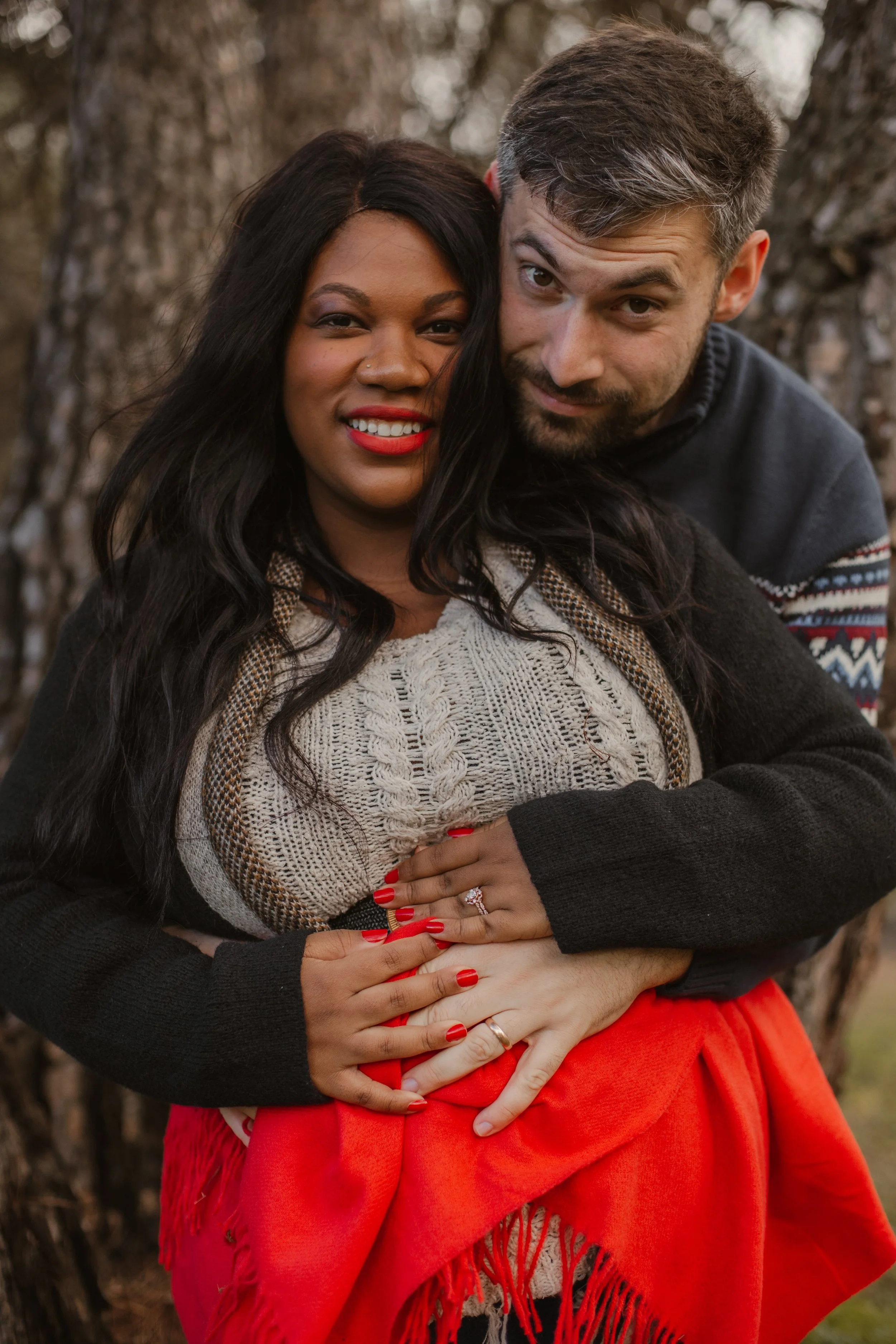 A couple embracing outdoors, with a woman in a cream and brown sweater and red skirt, and a man in a dark sweater with patterned sleeves. They are standing in front of tree trunks, with the woman showing her engagement ring and smiling, while the man