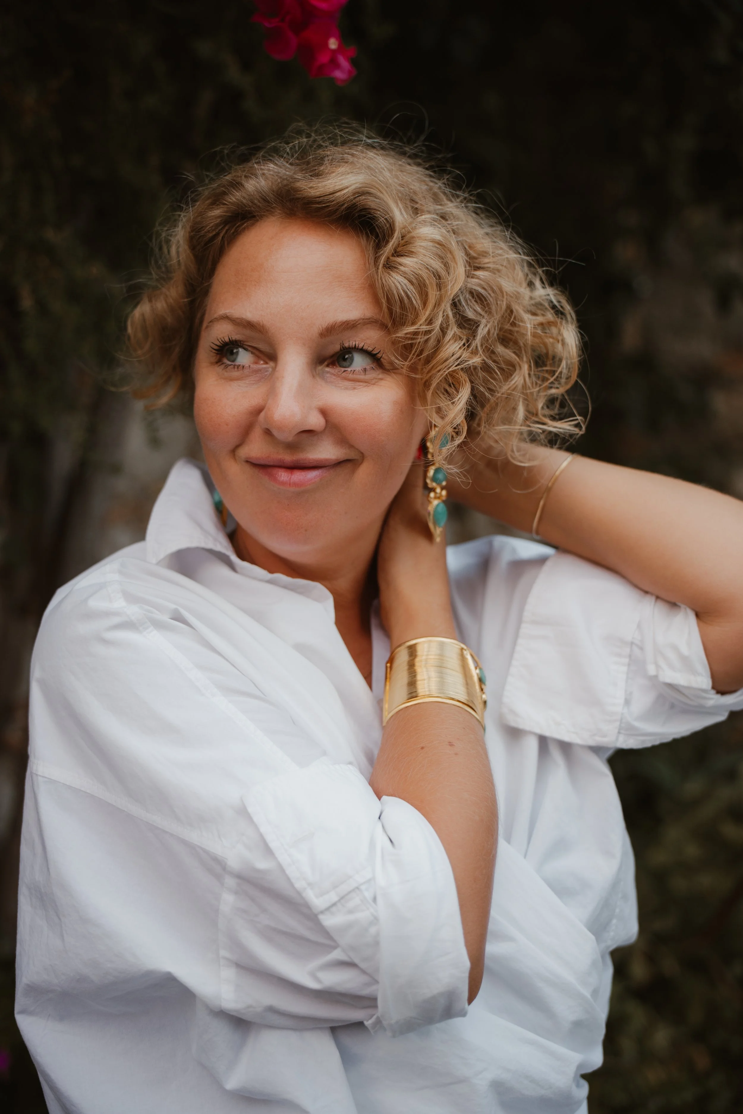 A young woman with long brown hair smiling while resting her head on her hand, sitting on the floor in front of a black chair, wearing a white shirt and blue jeans.