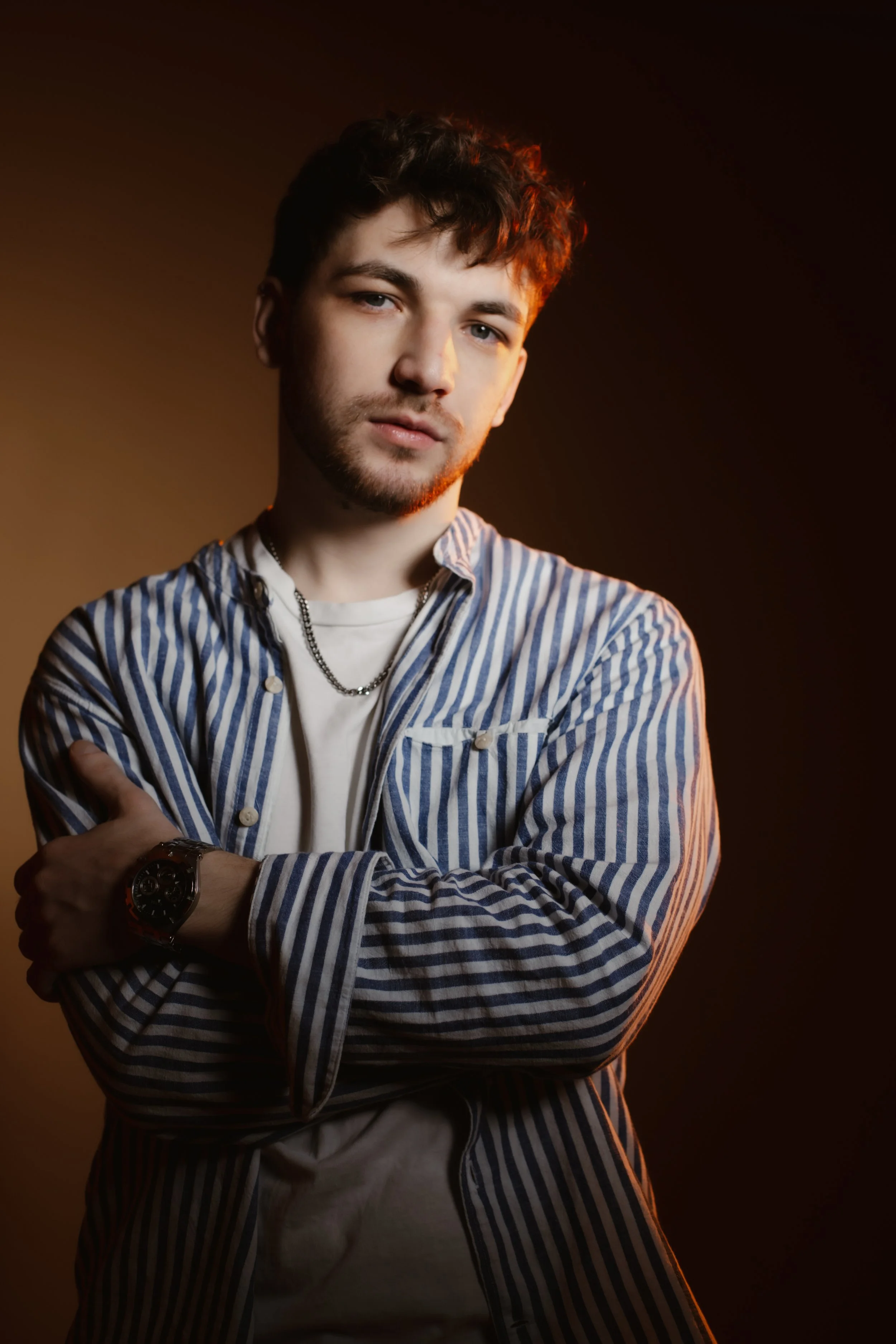A young man with dark hair and a beard wearing a striped shirt and a watch, standing with arms crossed against a dark background.