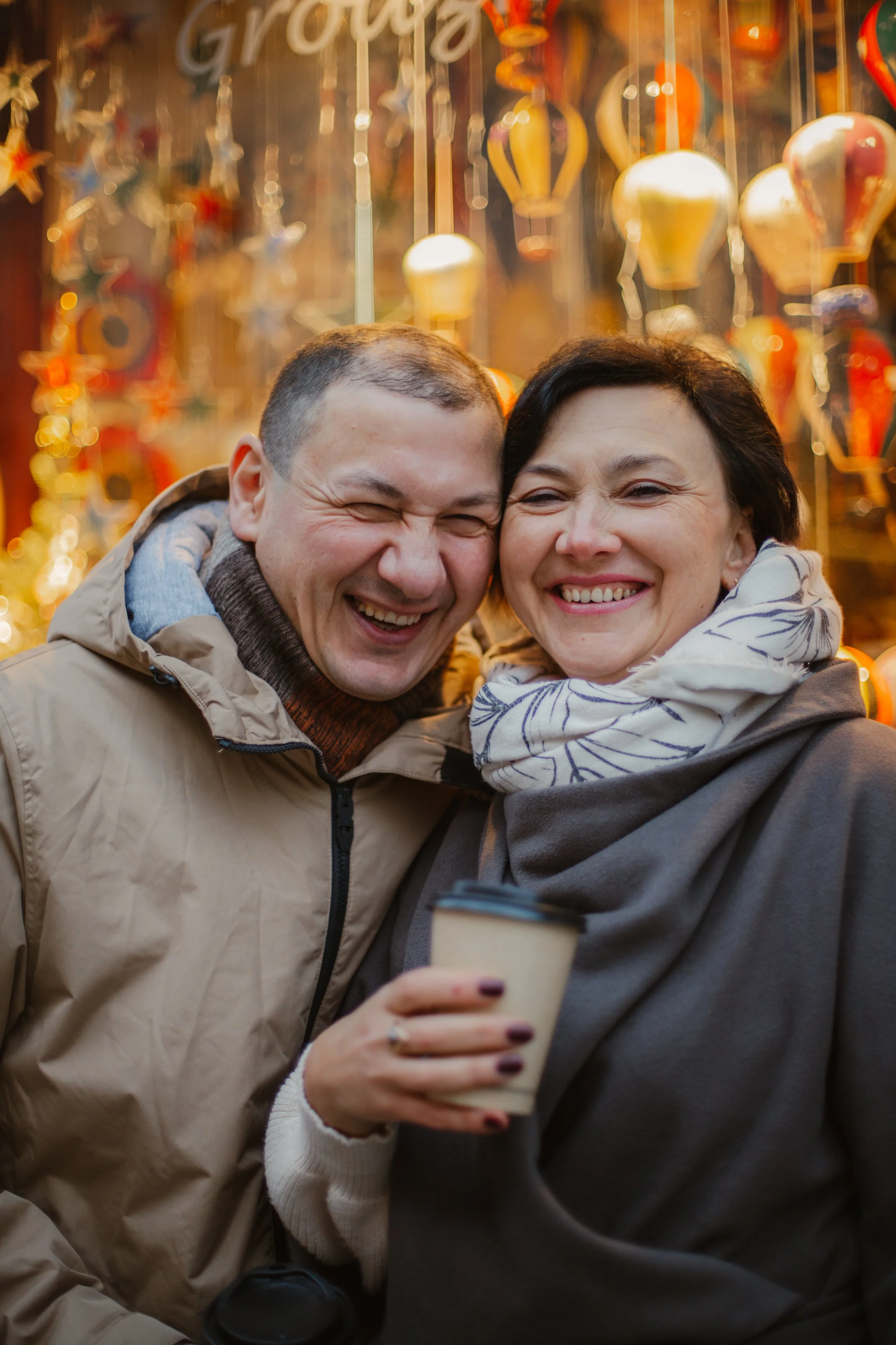 Happy couple at a holiday market, smiling, surrounded by hanging ornaments, one holding a coffee cup.