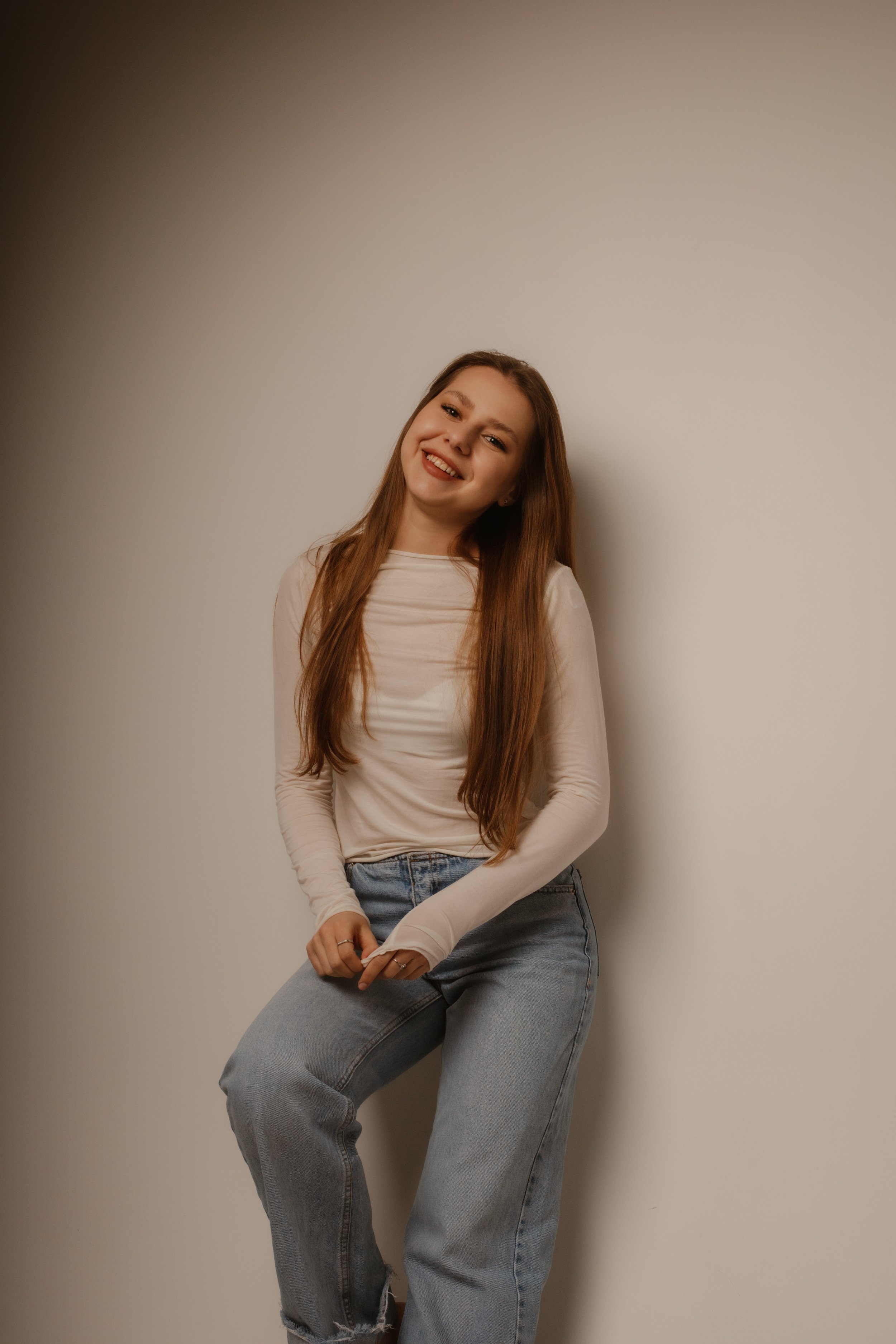 Young woman with long red hair smiling and leaning against a plain light-colored wall.