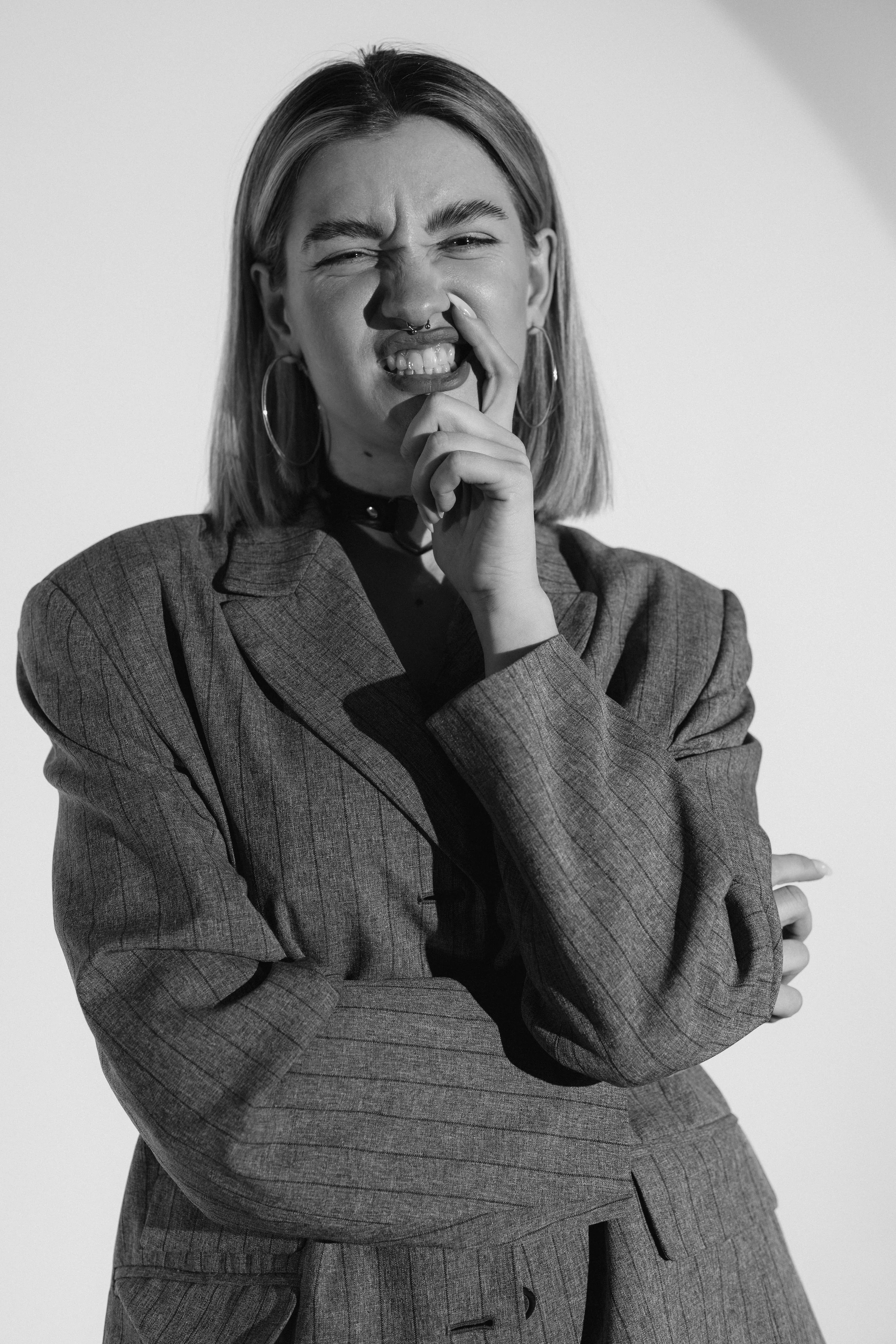 Black and white photo of a woman with shoulder-length hair, wearing a pinstripe blazer, making a puzzled or annoyed facial expression with one eye slightly squinted, and her hand touching her nose.