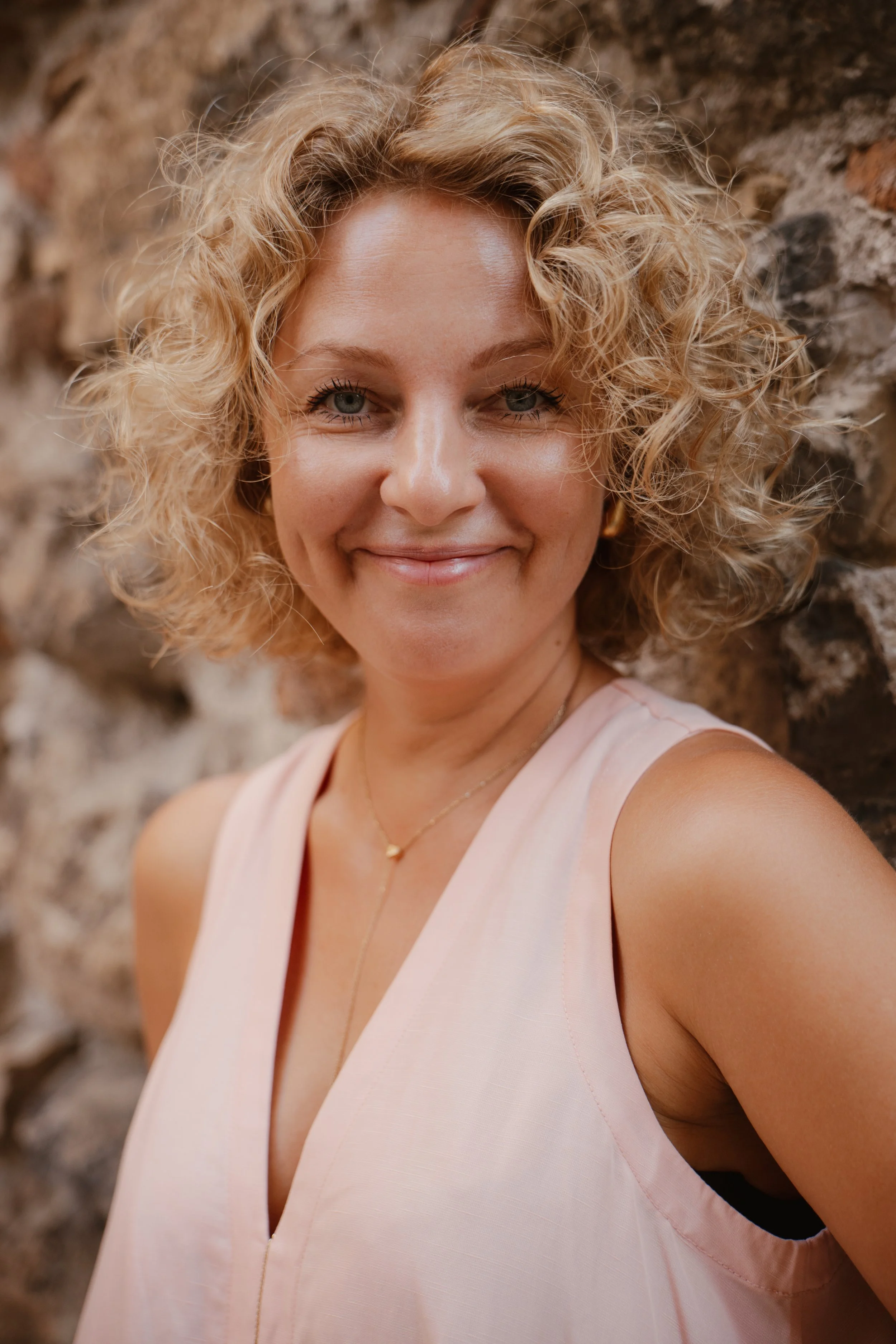 A woman with curly blonde hair smiling at the camera, standing in front of a stone wall.
