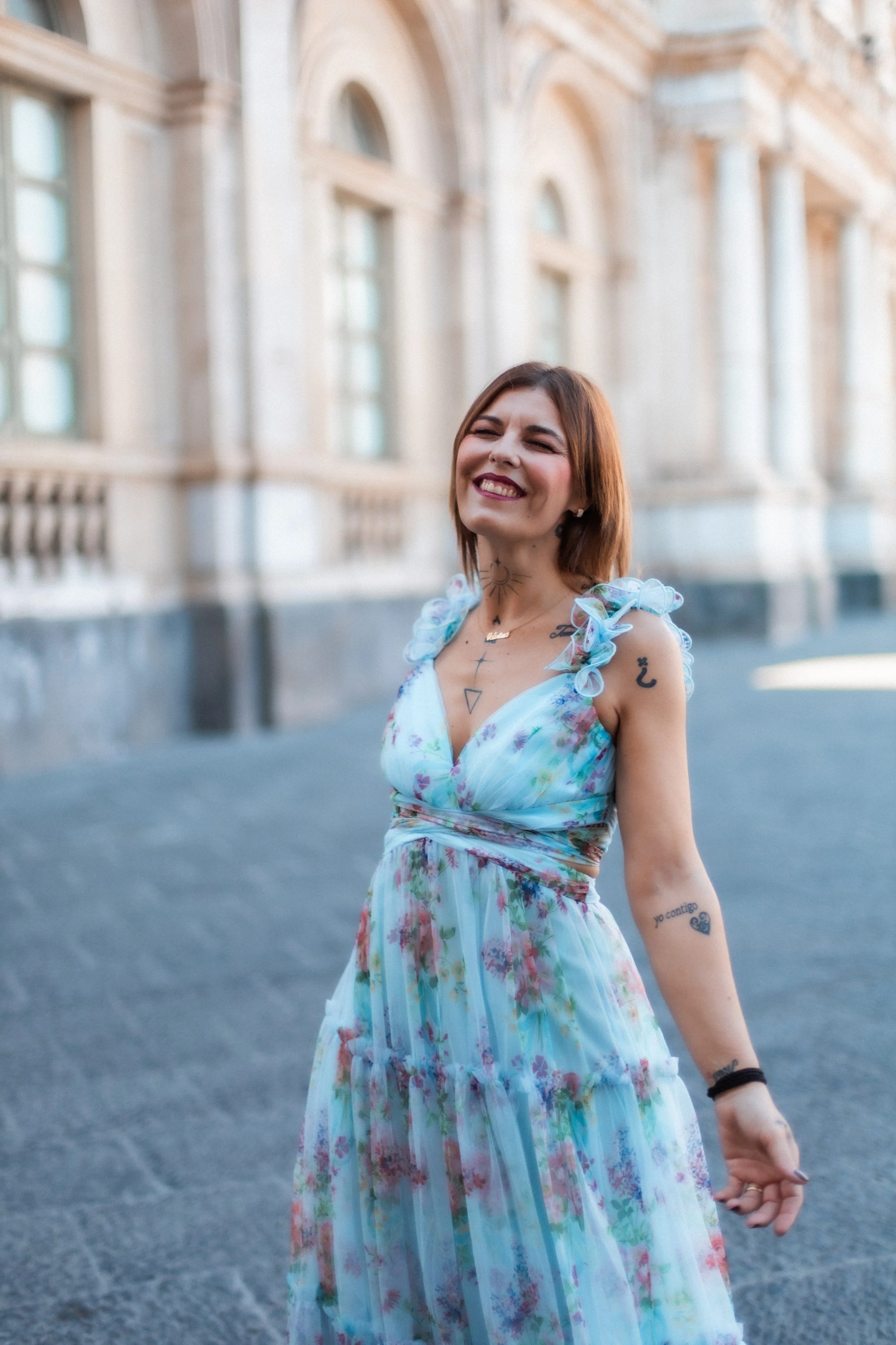 A woman with tattoos wearing a floral dress, smiling, in front of a historic building.