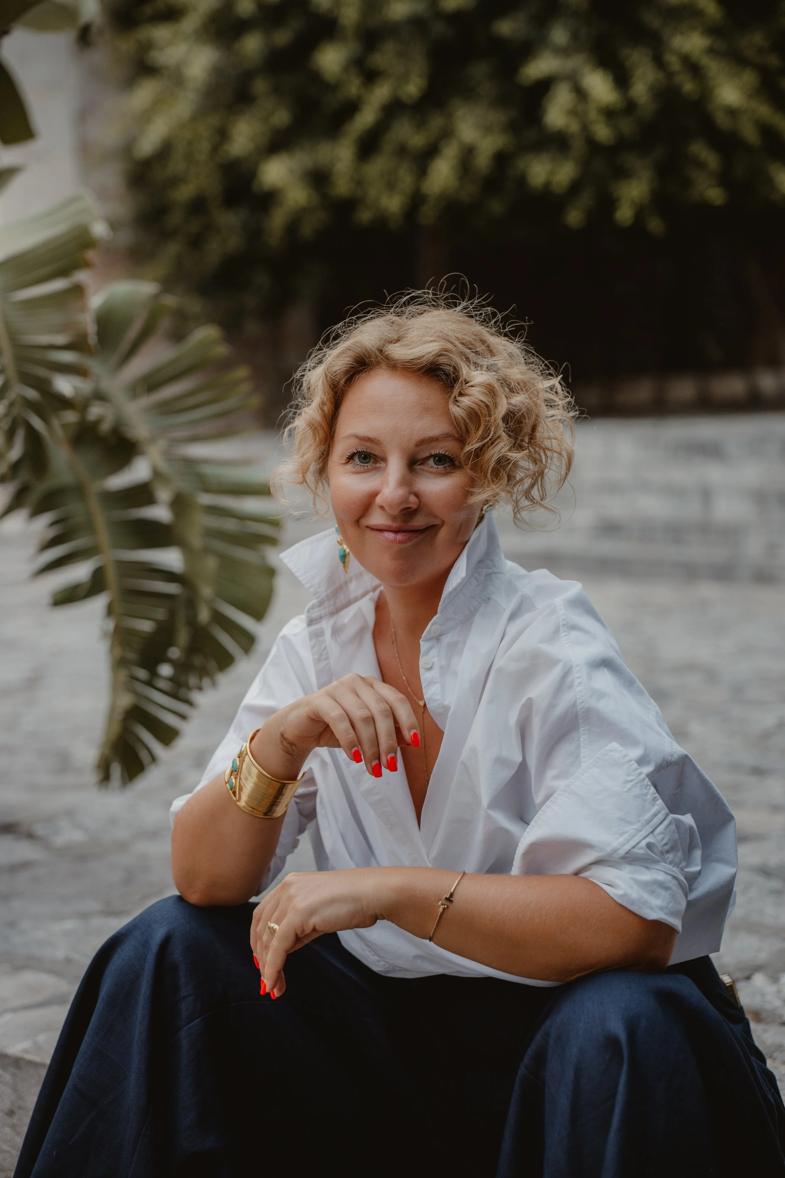 A woman with short curly blond hair wearing a white shirt, dark pants, and colorful jewelry, sitting outdoors near a body of water with greenery in the background.